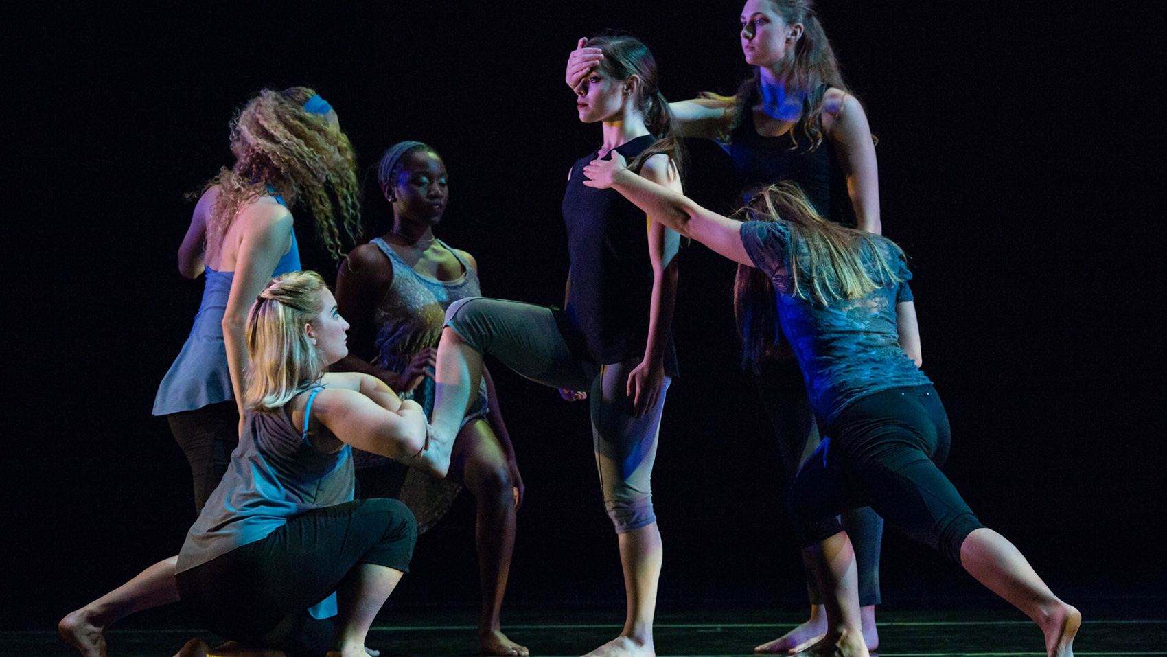 One female dancer stands still in the middle, she rests her arms by her side and raises one leg. Behind her one female dancer raises her arm to cover one of the center dancer's eyes. Another dancer lunges forward as she raises one hand to cover the center dancer's heart. Three other dancers are grouped in front of the center dancer, each with varying poses.