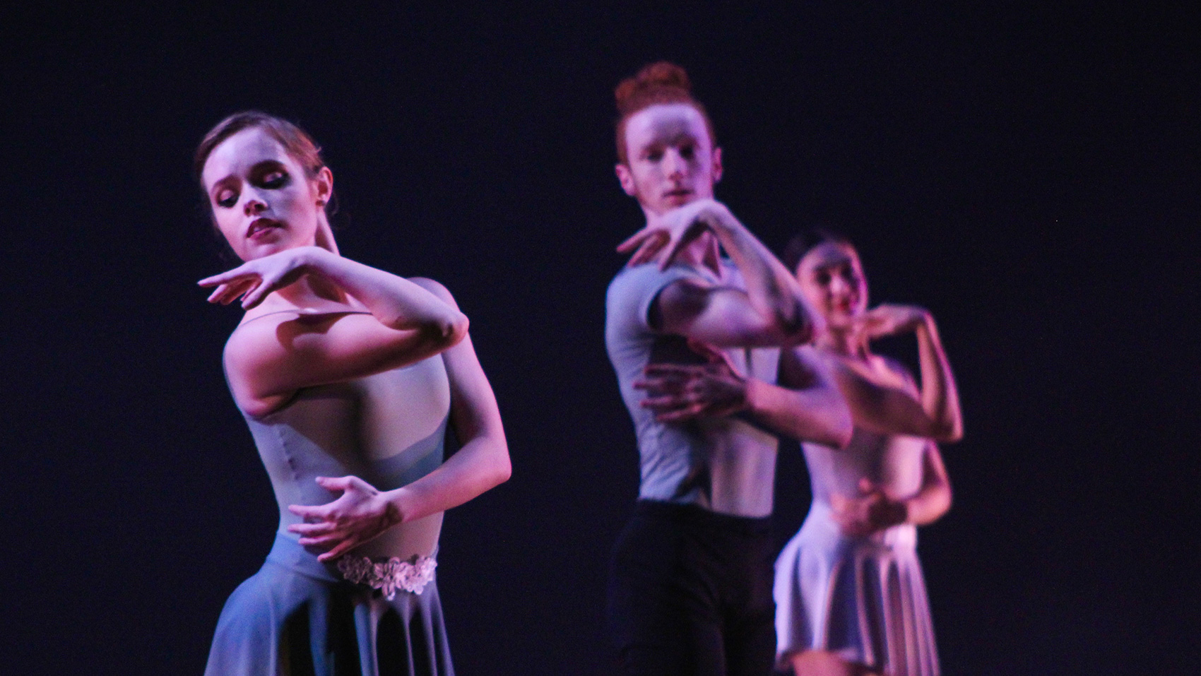 Three ballet dancers hold the same pose: they pose slightly to the side, they look over their right shoulder and raise their right arm to the front, lifting their hand to graze their chin. Their rest their other hand on the right side of their stomach.
