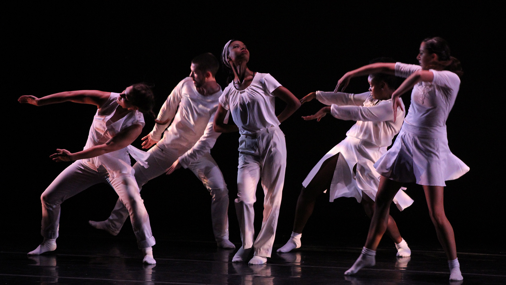 One Black female dancer stands in the center, her hands behind her back. She looks upwards. Four other dancers beside and behind her dance the same pose, they stretch their arms and turn their faces in the direction opposite to the center dancer.