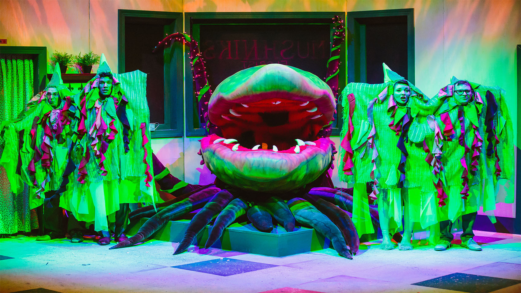 A giant bulbous plant with sharp teeth has its mouth open while being surrounded by characters draped in fabric to look as though they are assimilated with the plant. 