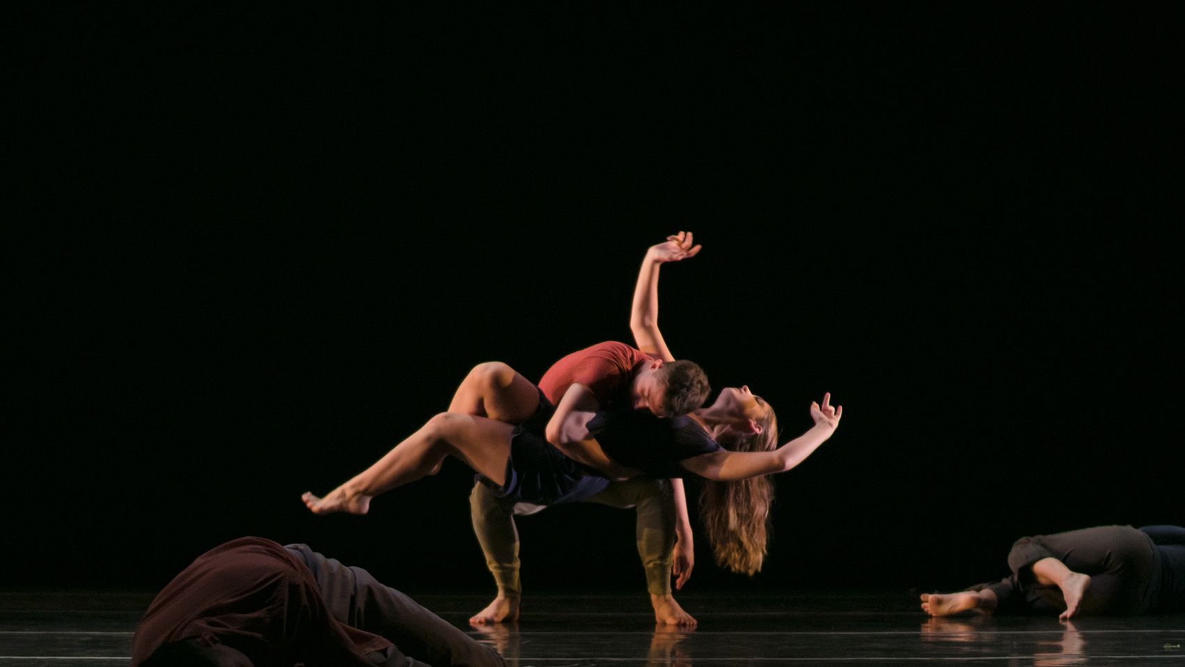 A male dancer crouches his body as he embraces and holds a female dancer with one arm, his other arm trailing to his side. The female dancer lays in his embrace, her body parallel to the ground. She moves her arms gracefully through the air.