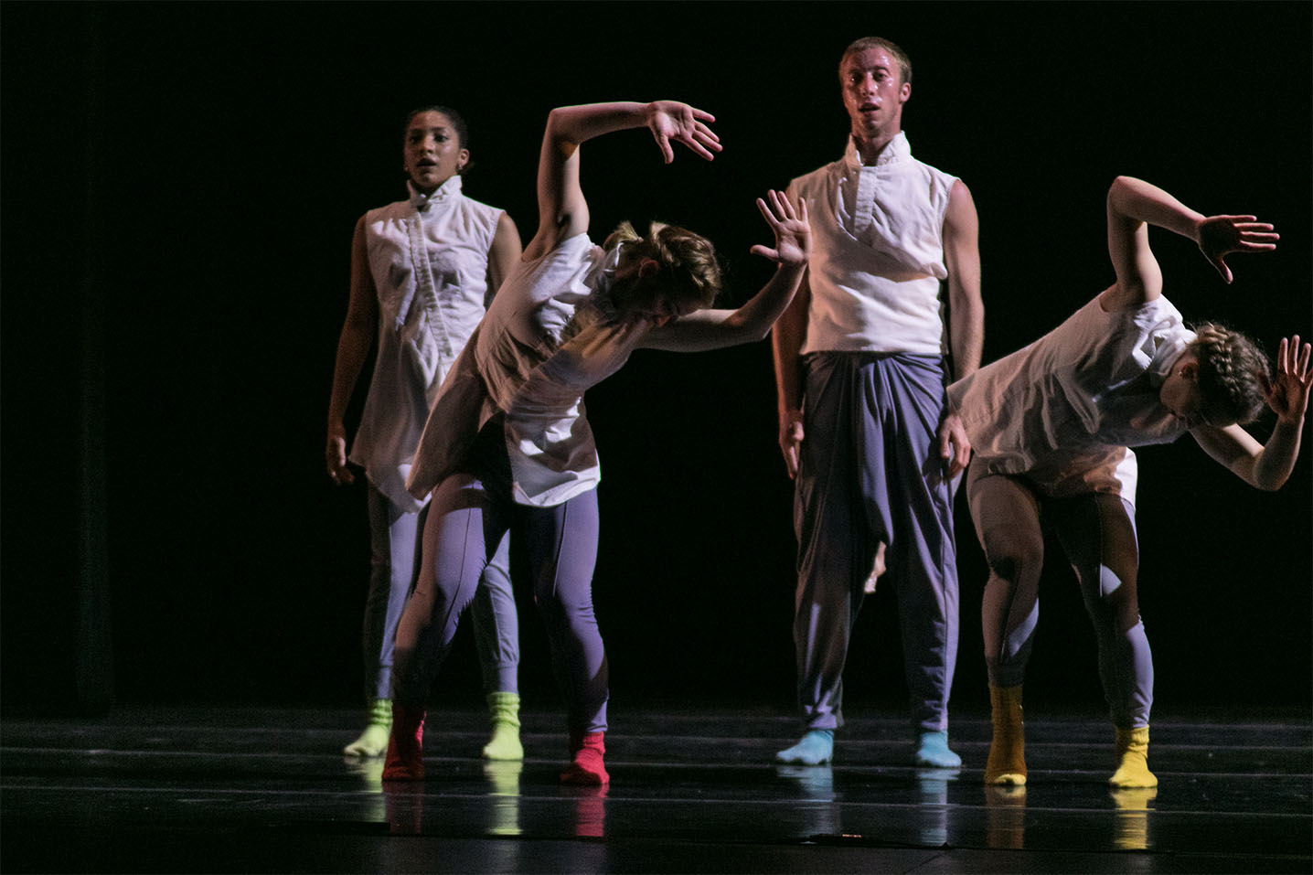 Two female dancers in the front bend their torso to the right side, their arms raised above their heads. Behind them, one female dancer and one male dancer stand skill, arms resting at their sides. All dancers are wearing white tops, purple-gray pants, and bright colorful socks.