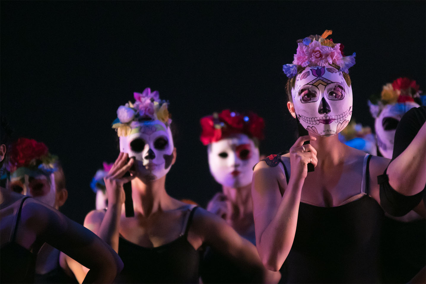 Female dancers all wearing black spaghetti strap tank tops hold white decorated masks over their faces. All wear brightly colored flowers at the front of their heads. 