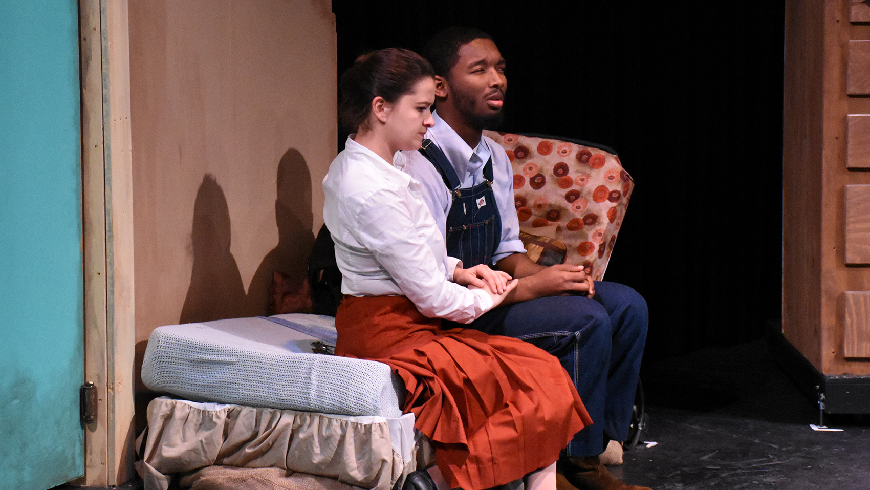 A woman and a man sit on what looks like a sofa bed, both with concerned faces. The woman places her hands on the man's arm, as if to comfort him.