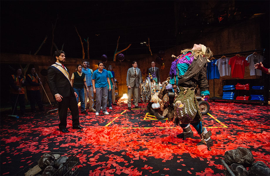 Twelve characters stand watching a blindfolded female character that is wearing brightly colored clothes, holding many bags, and is leaning back to face the ceiling.