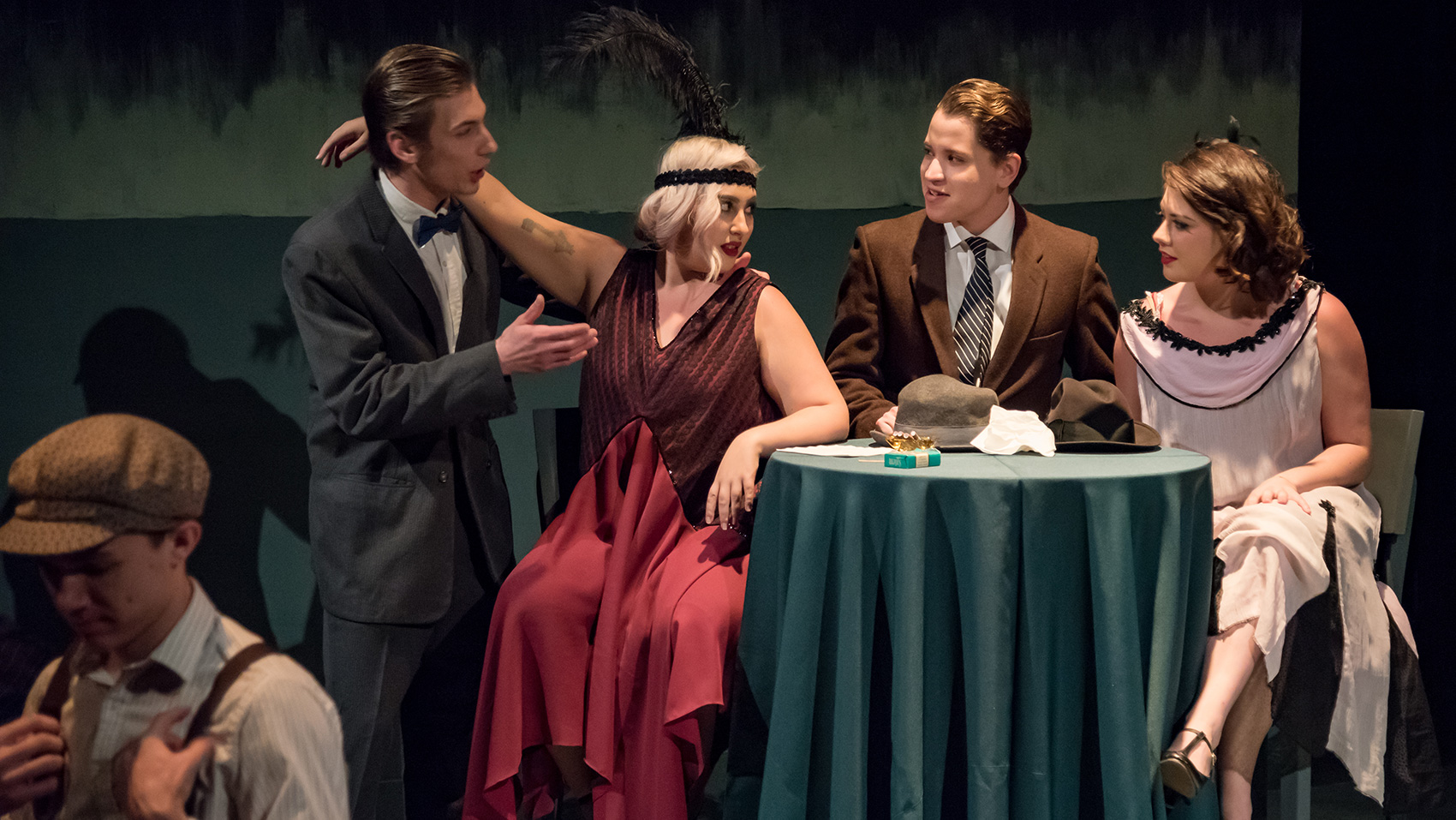 Two women and a man sit together at a table, one woman with her arm draped over the shoulder of a man who is standing and talking to them. All of them are dressed formally, the women wearing nice dresses, one man wearing a tie and the other a bowtie.