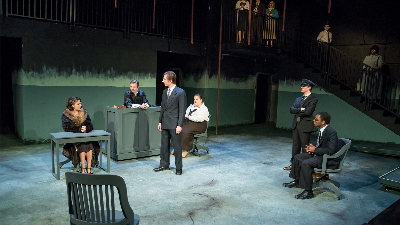 The stage  has been converted into a courthouse. The woman sits at a table, interrogated by a lawyer. The judge and other people look on.