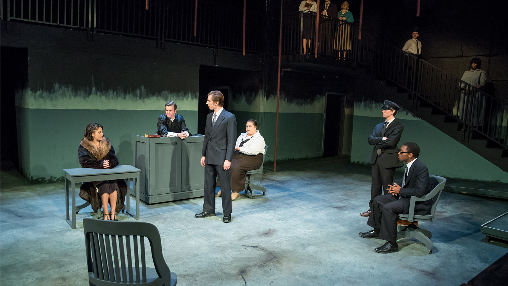 The stage has been converted into a courthouse. The woman sits at a table, interrogated by a lawyer. The judge and other people look on.