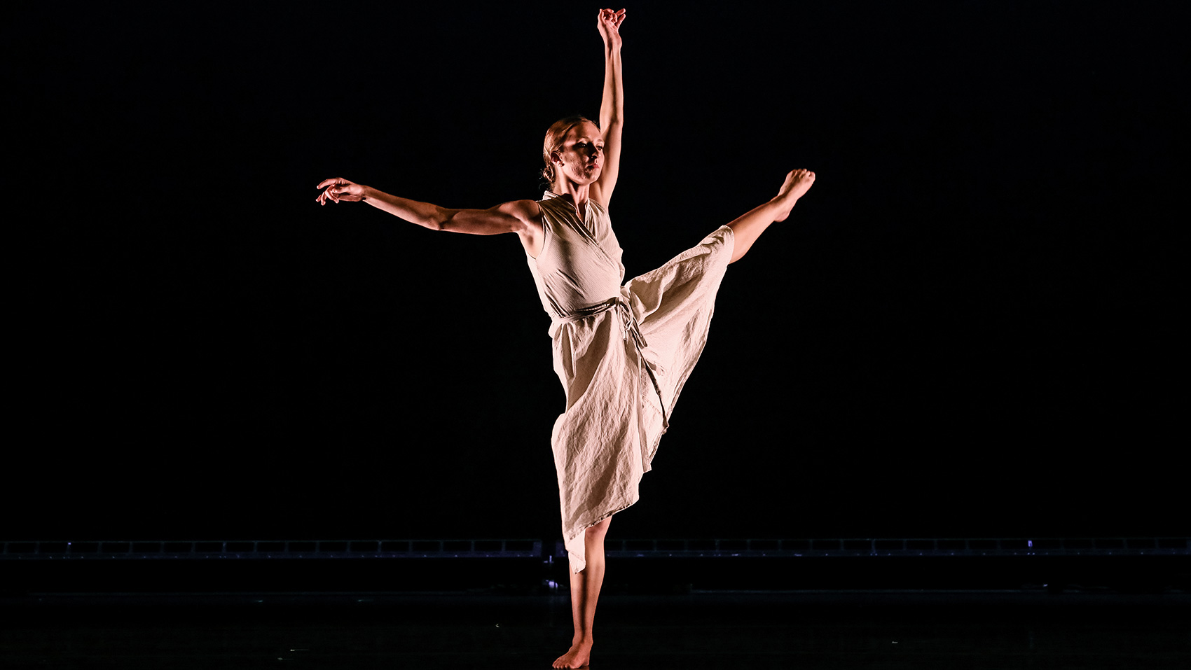 One woman dancer extends her arms, one above her head, the other to her side. She extends one leg high to her side.