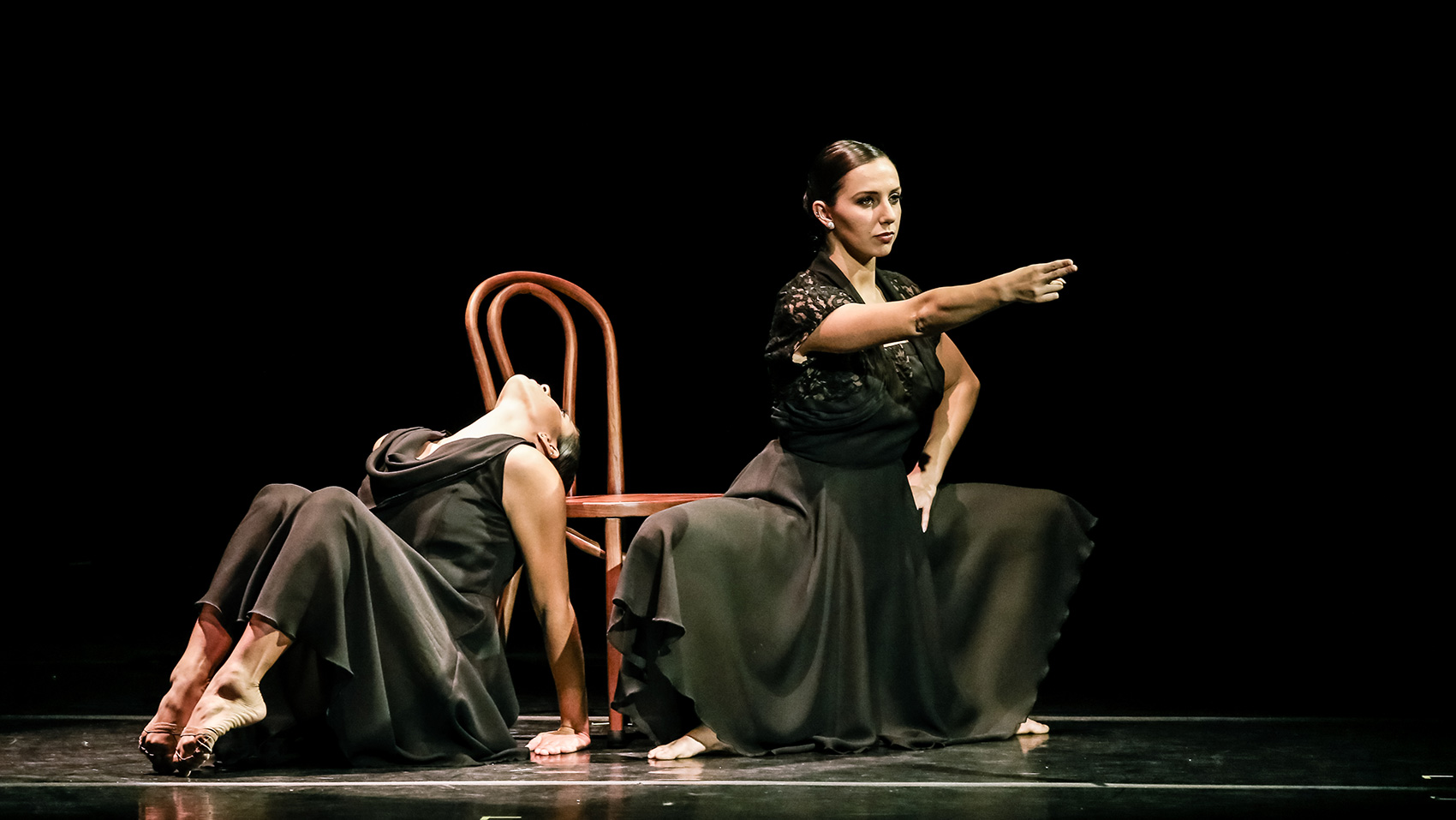 Two women dancers. One of them squats in front of a chair, one hand on her hip and the other hand stretched straight in front of her, the index finger and middle finger pointing ahead. The other woman arches her back, her face to the ceiling, her feet are pulled together with her toes on pointe.