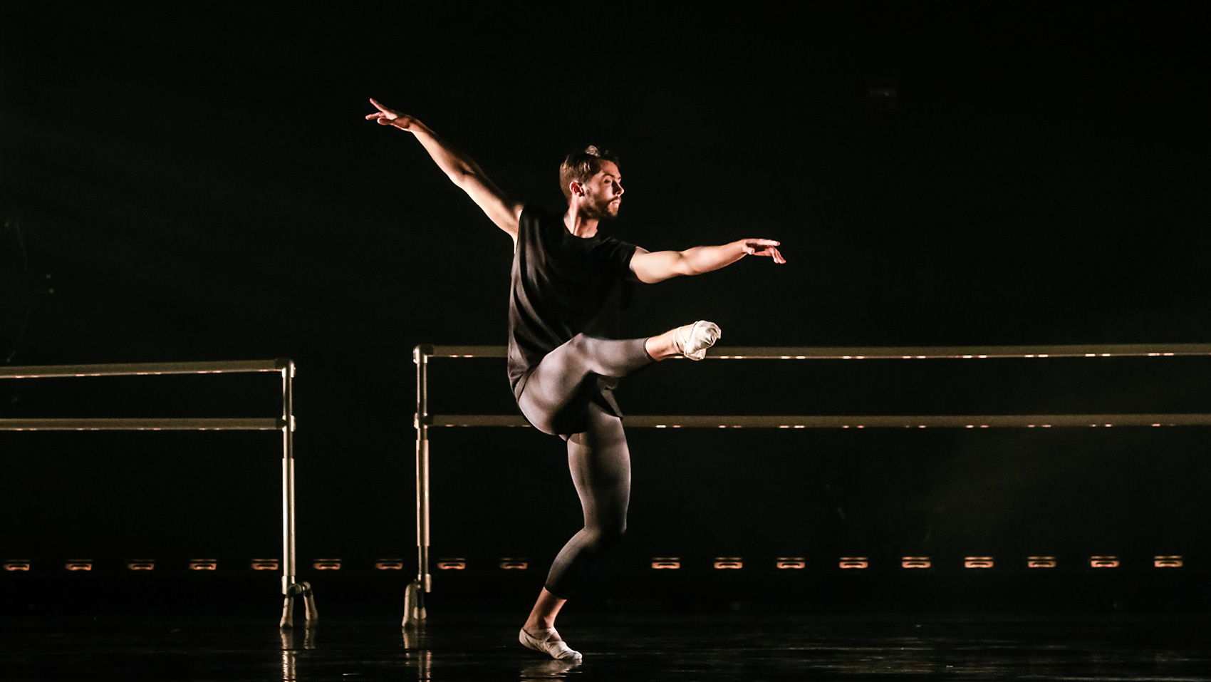 One male dancer dances on pointe, both arms extended gracefully.