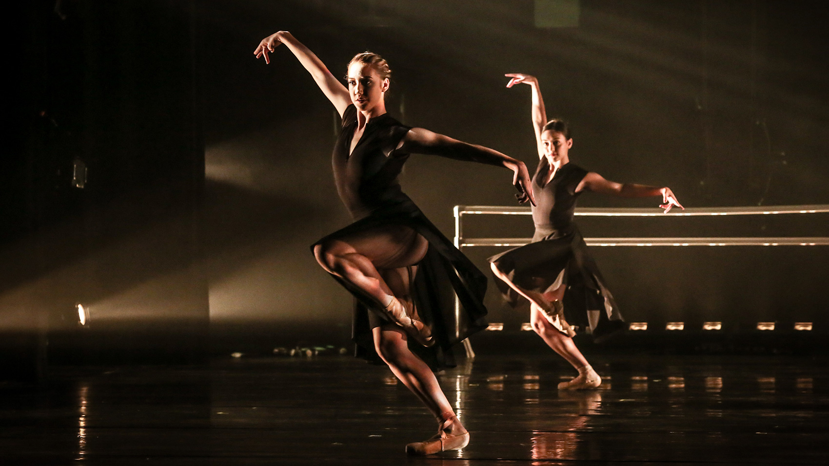 Two women dancers in the same dance pose: One arm extended above their head, the other extended to their side. One leg raised with the knee bent inward, toes on pointe. The other foot is firmly on the ground to support their movement.