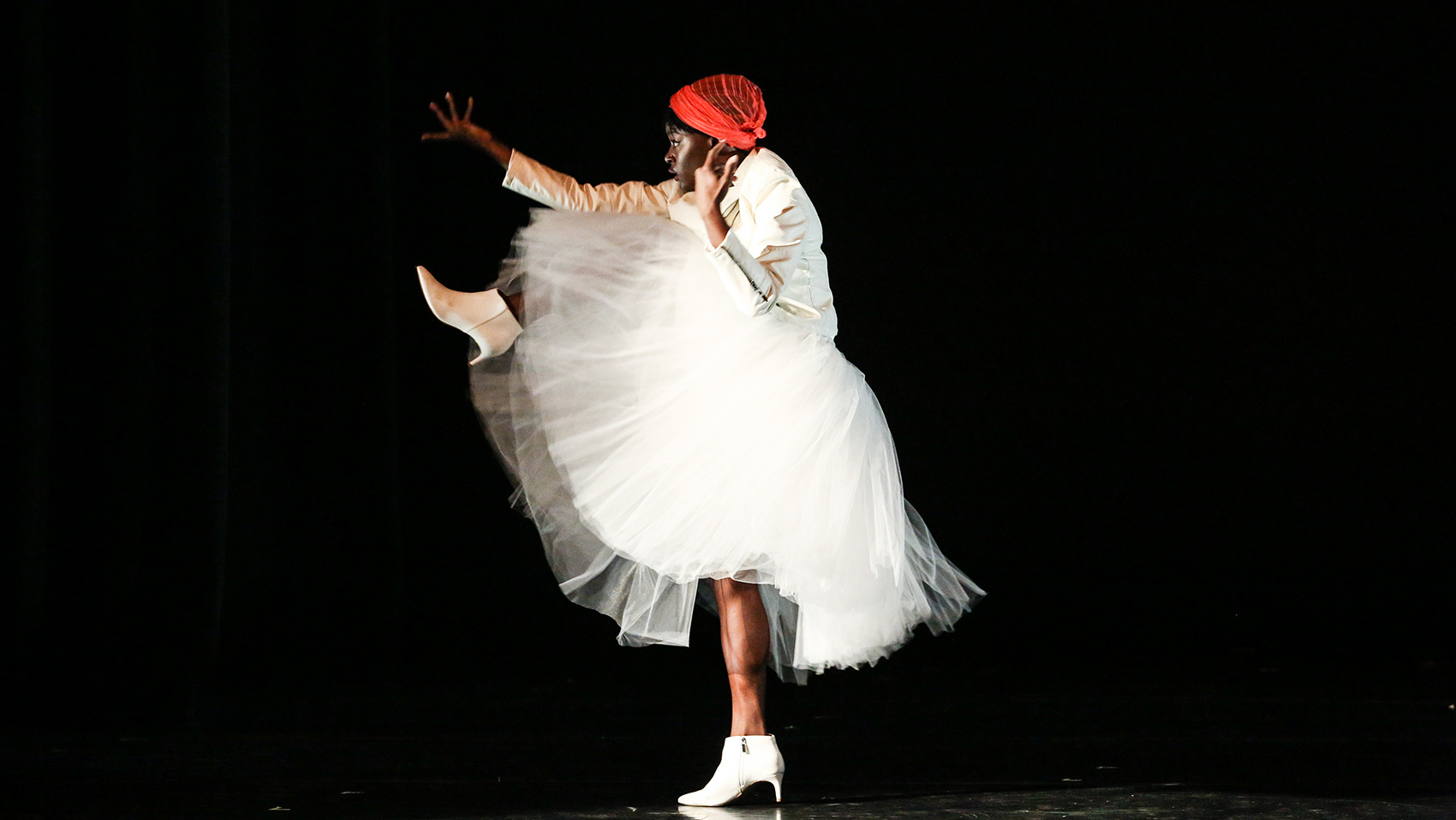 A Black woman dancer wearing a white shirt, white tutu skirt, and white ankle boots. The picture captures her mid-dance-pose from the side. she is in profile. She raises one leg to her chest, stretches one hand in front with her palm facing out.