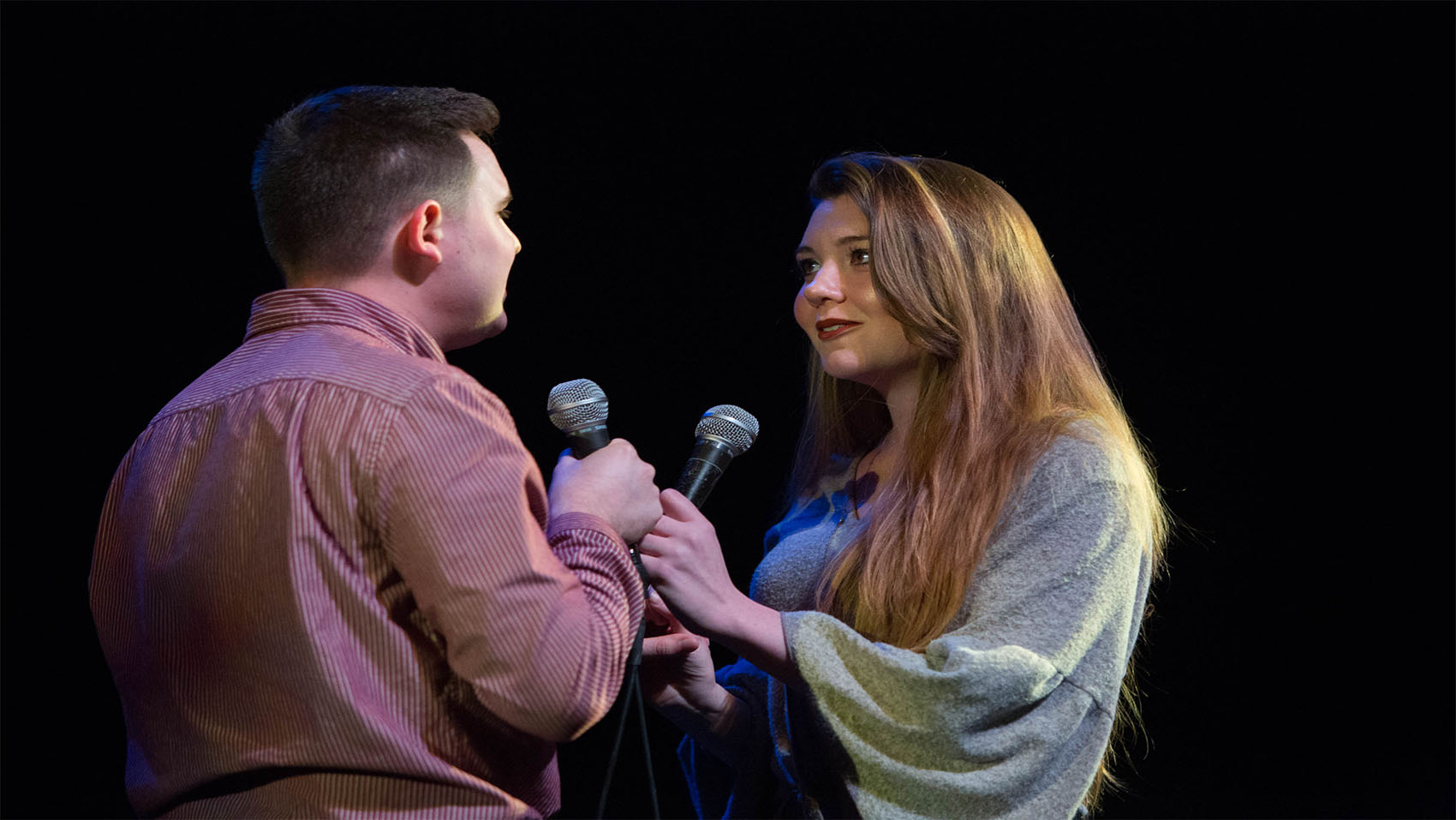 A young man and woman are embraced, looking at each other in adoration, microphones facing each other.