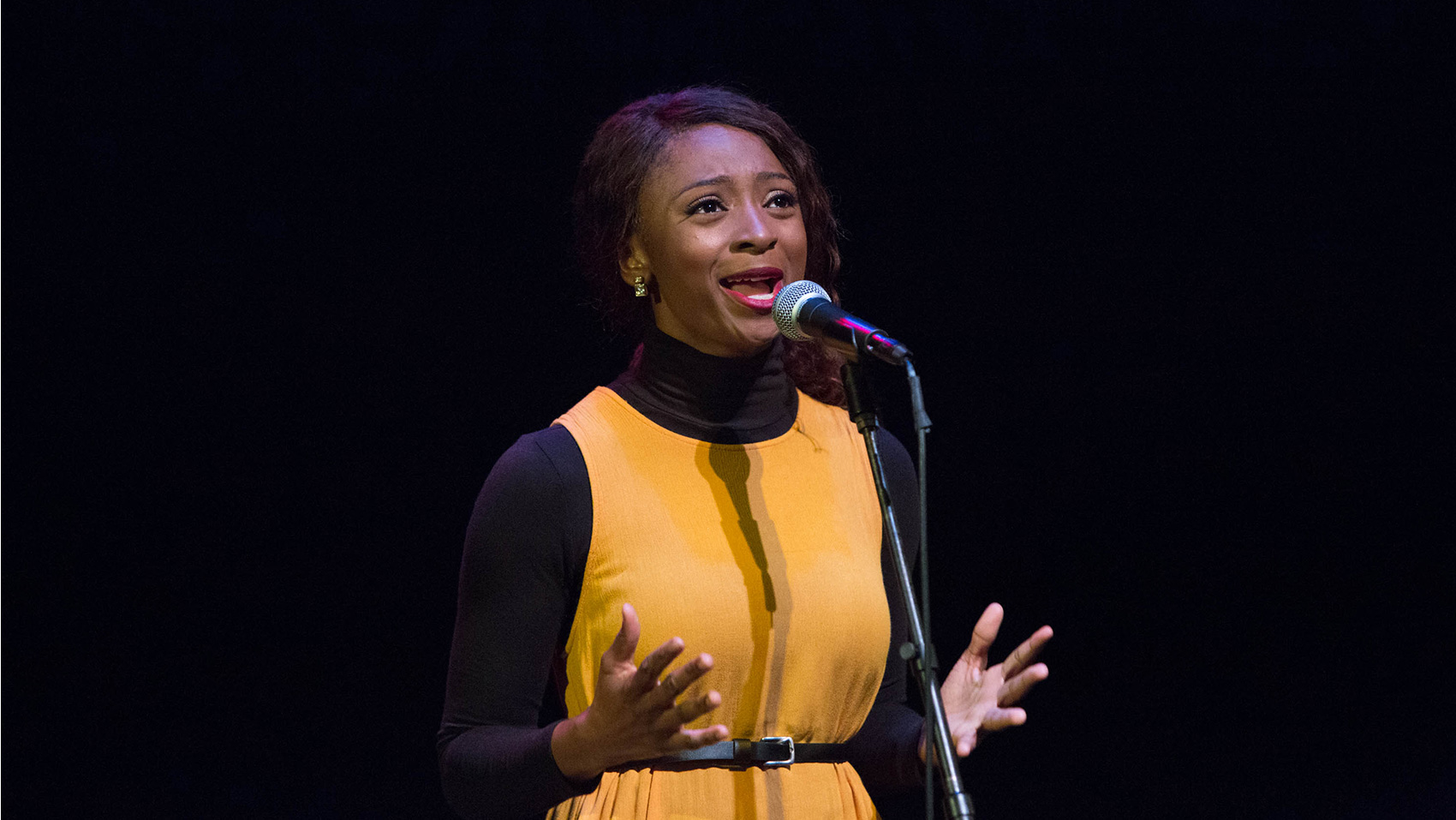 A young woman looks upward while singing,her hands splayed out. Her face is masked with adoration.