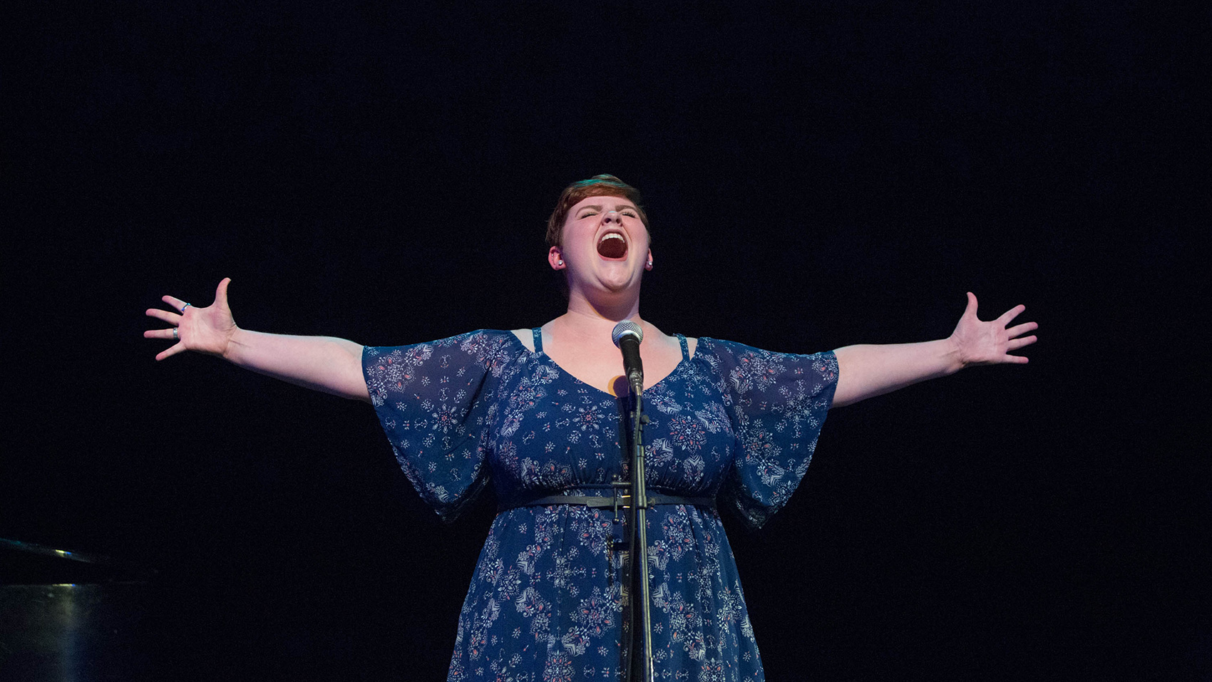 A woman sings loudly, her arms outstretched on either side of her as she does.