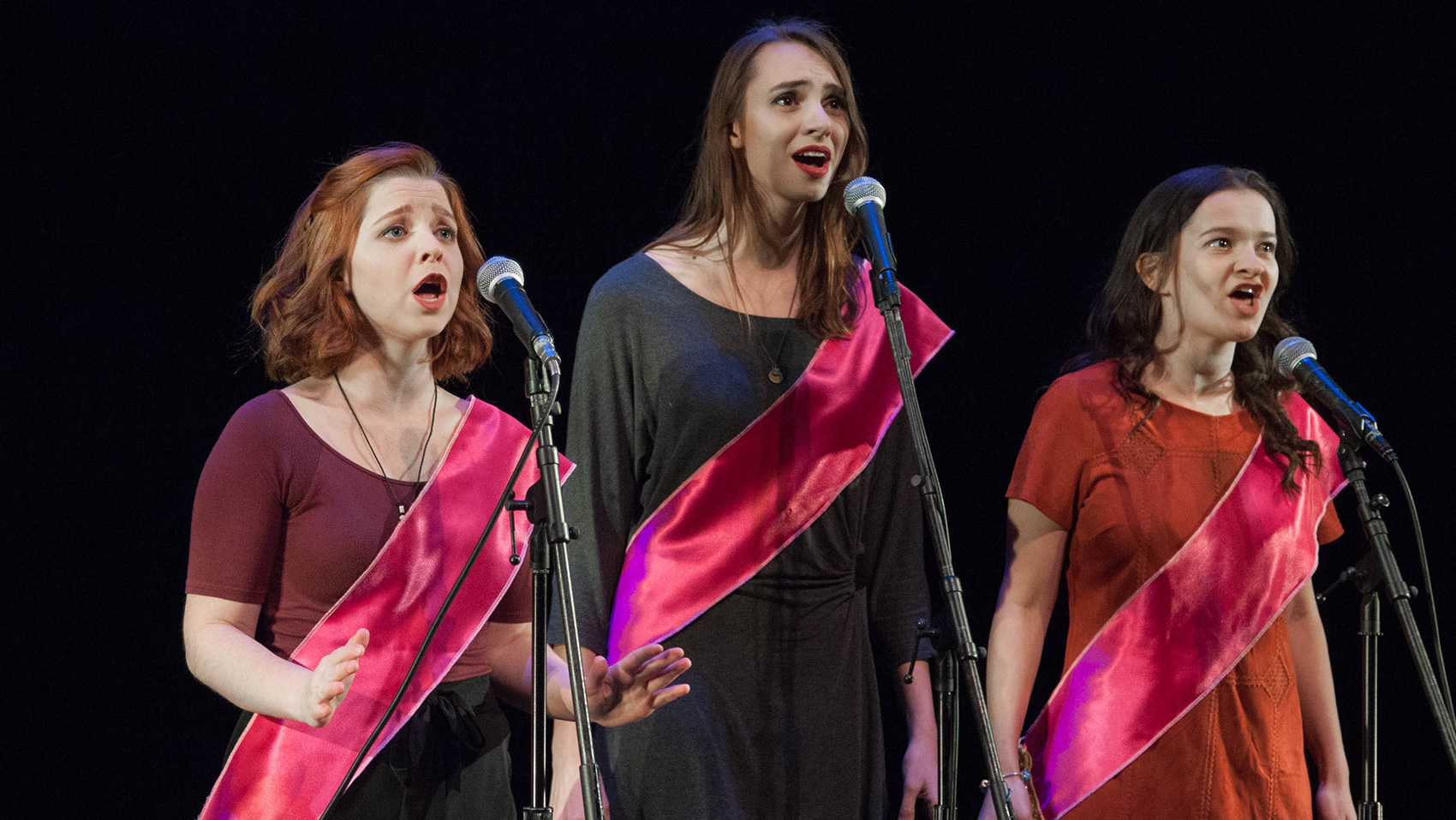 Three women stand in a group singing, each wearing a silky red sash across their body. The woman on the far left has a face of slight worry, the middle woman seems hopeful and the woman on the right has a face of excitement/tenacity