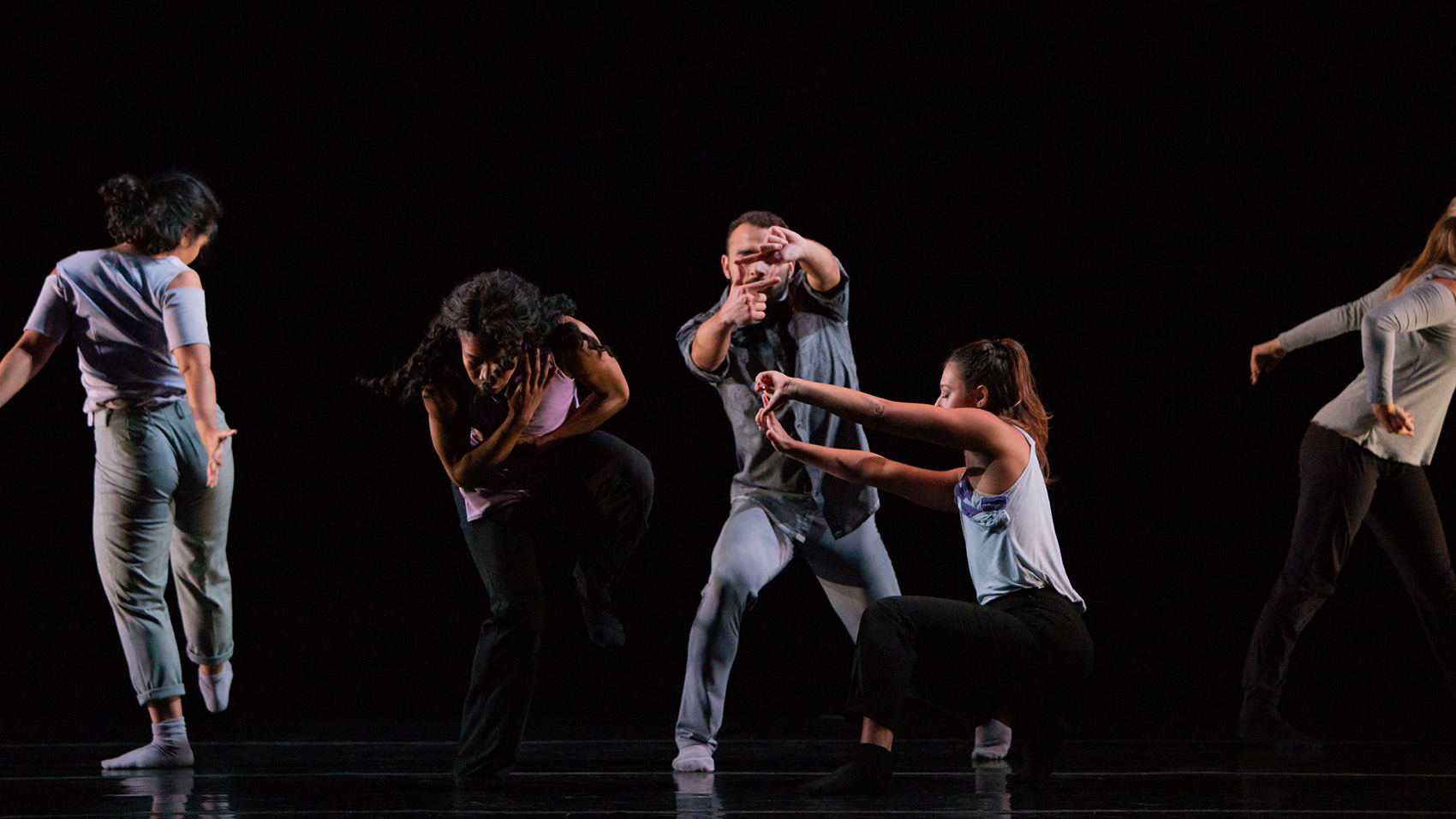 Five dancers on stage, all with different poses. One of them is bending her waist to learn forward, one leg raised. One hand is on her stomach, the other is on her chest, she looks as if she is holding herself. Behind her, there is a male dancer whose hands are making the shape of a rectangle, as if he is taking a picture.