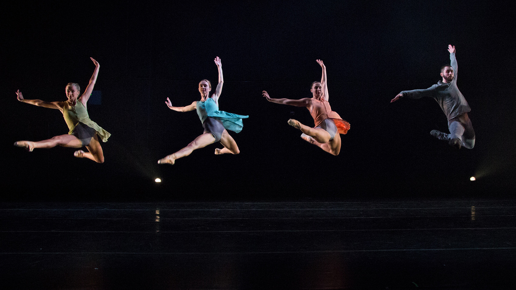 Five dancers in different colored clothes are in mid jump on stage.