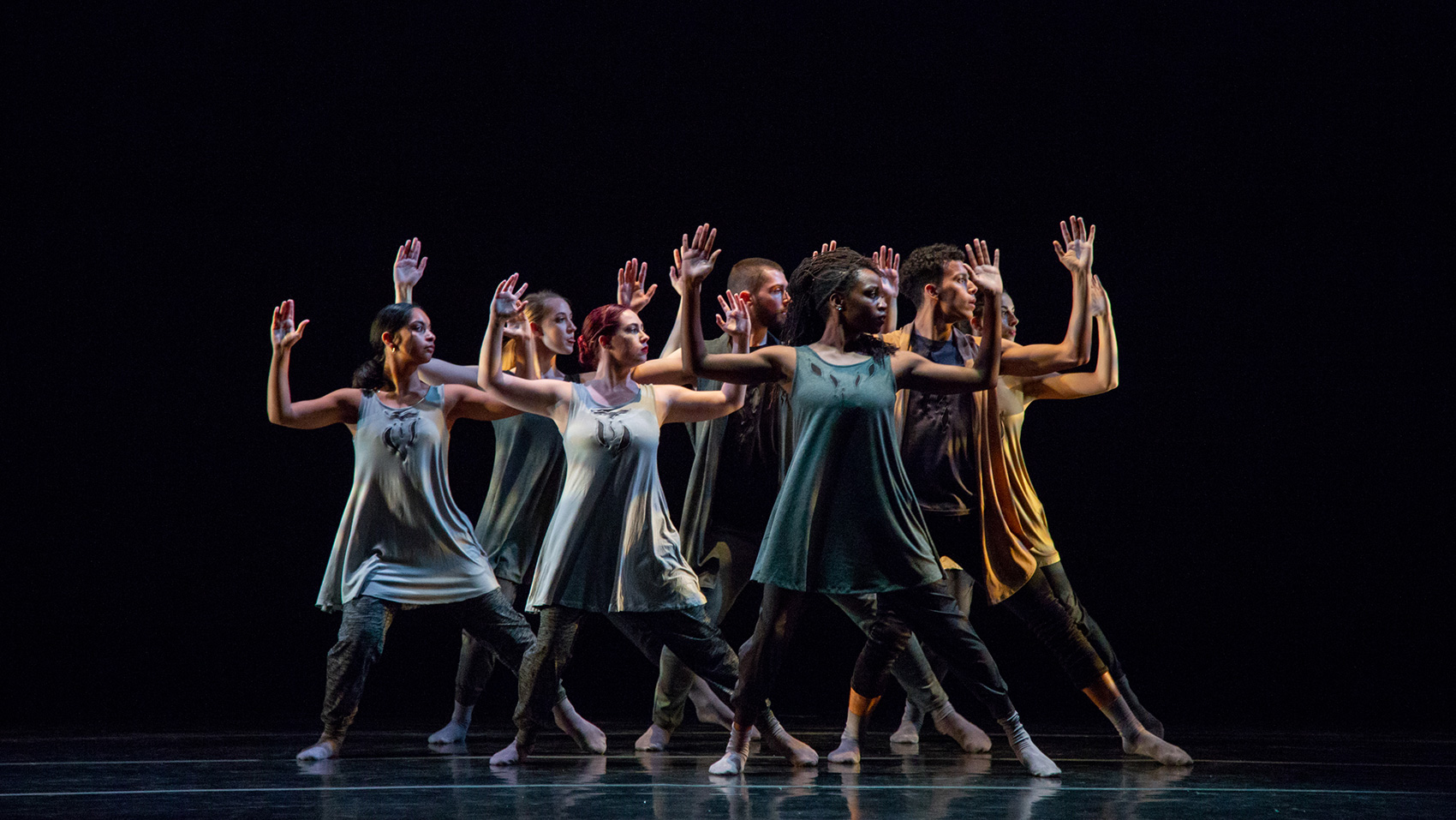 Seven dancers hold the same pose: their arms spread open side to side, elbows bent at 90 degree, palms open. They all turn their heads to the right side, and they stand with their legs spread apart.