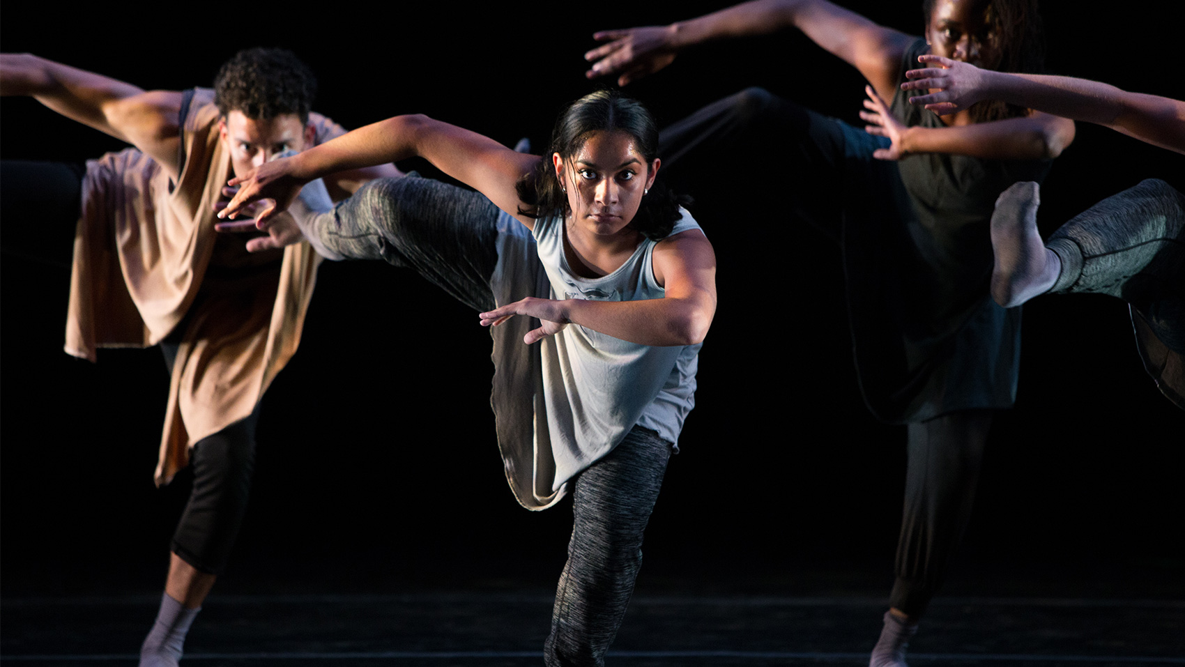 Three dancers stare straight ahead with a piercing stare. They hold the same pose: bent at the waist to lean forward, one arm spread behind them and one arm before them, as if they are swimming. They have also raised one leg behind them.