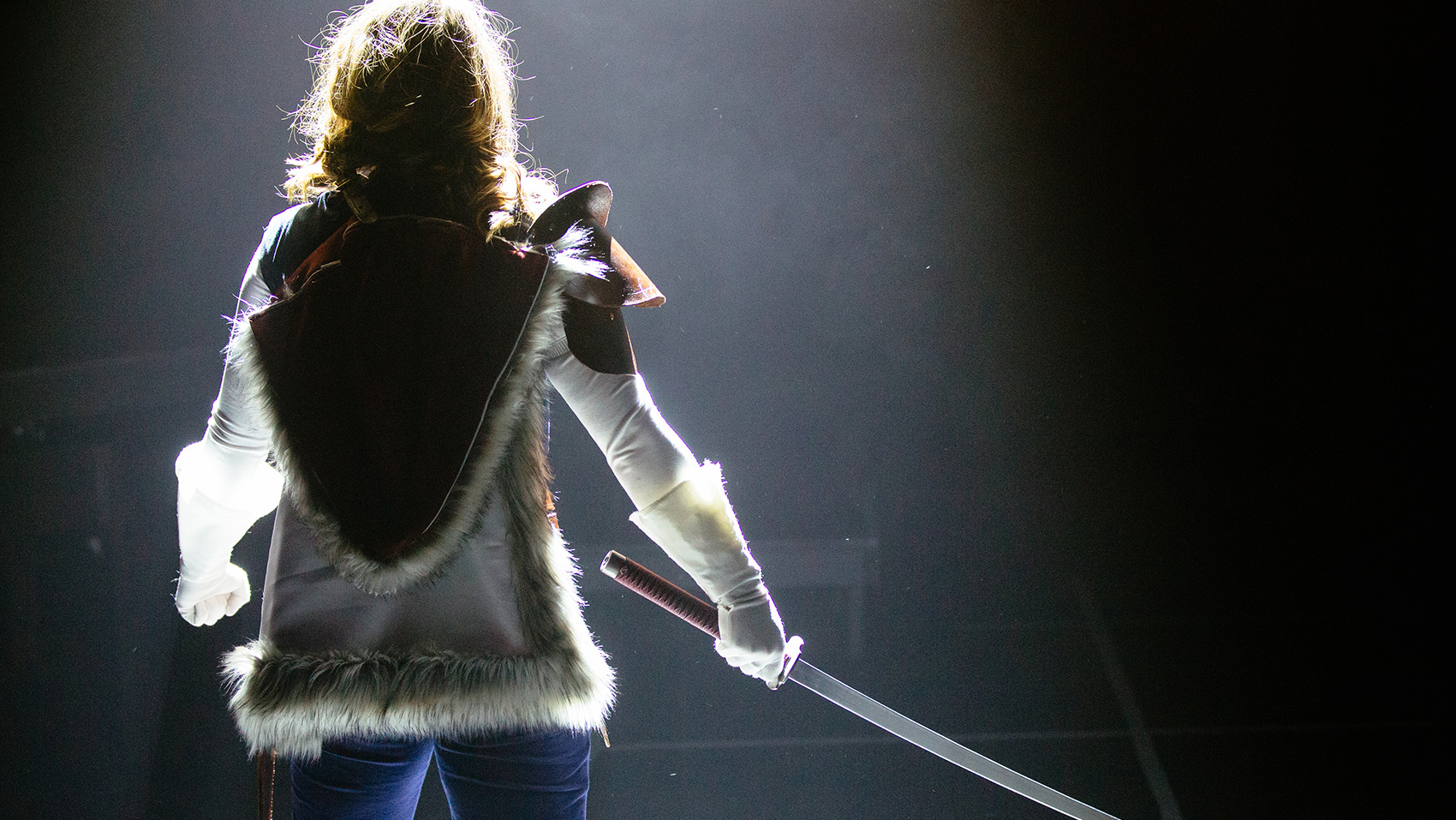 The back of a woman, in armor tensely holding a sword.