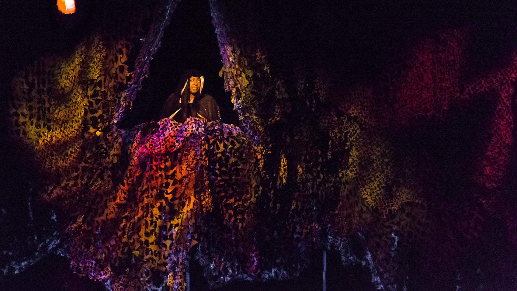 A woman stands at the opening of a cave/balcony, looking off into the distance, yellow-orange lighting cascading her face..