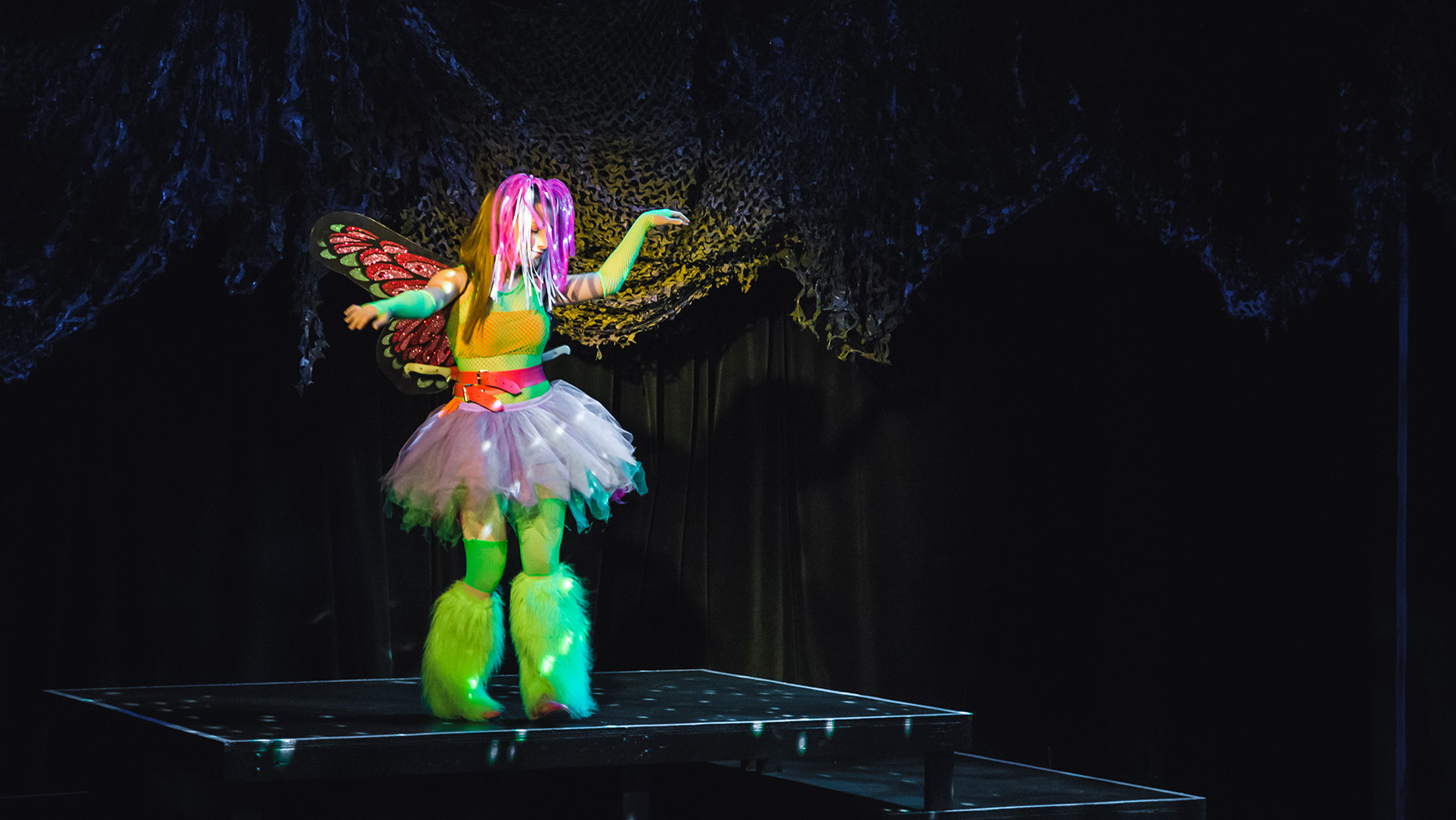 A woman decked in a tutu,fuzzy neon green boots, and butterfly wings stands on a platform, a look of grace on her face as she holds her arms out.