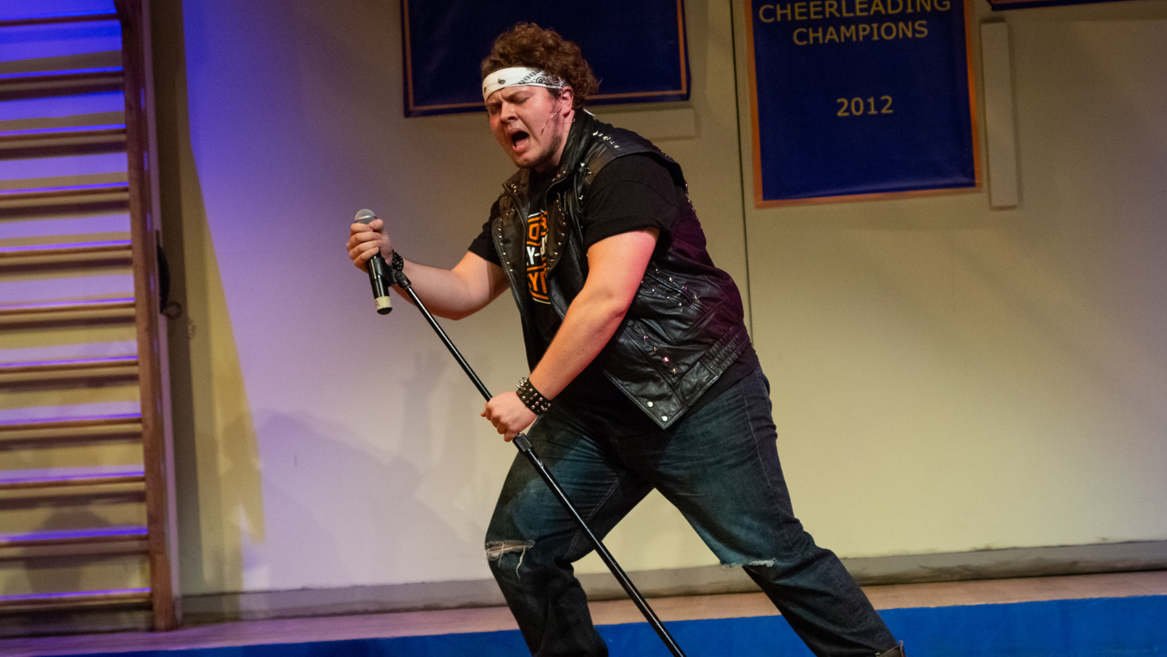 A man holds a microphone stand passionately as he sings into it. He wears a black leader vest over a black t-shirt and blue jeans, as well as a bandana on his forehead.