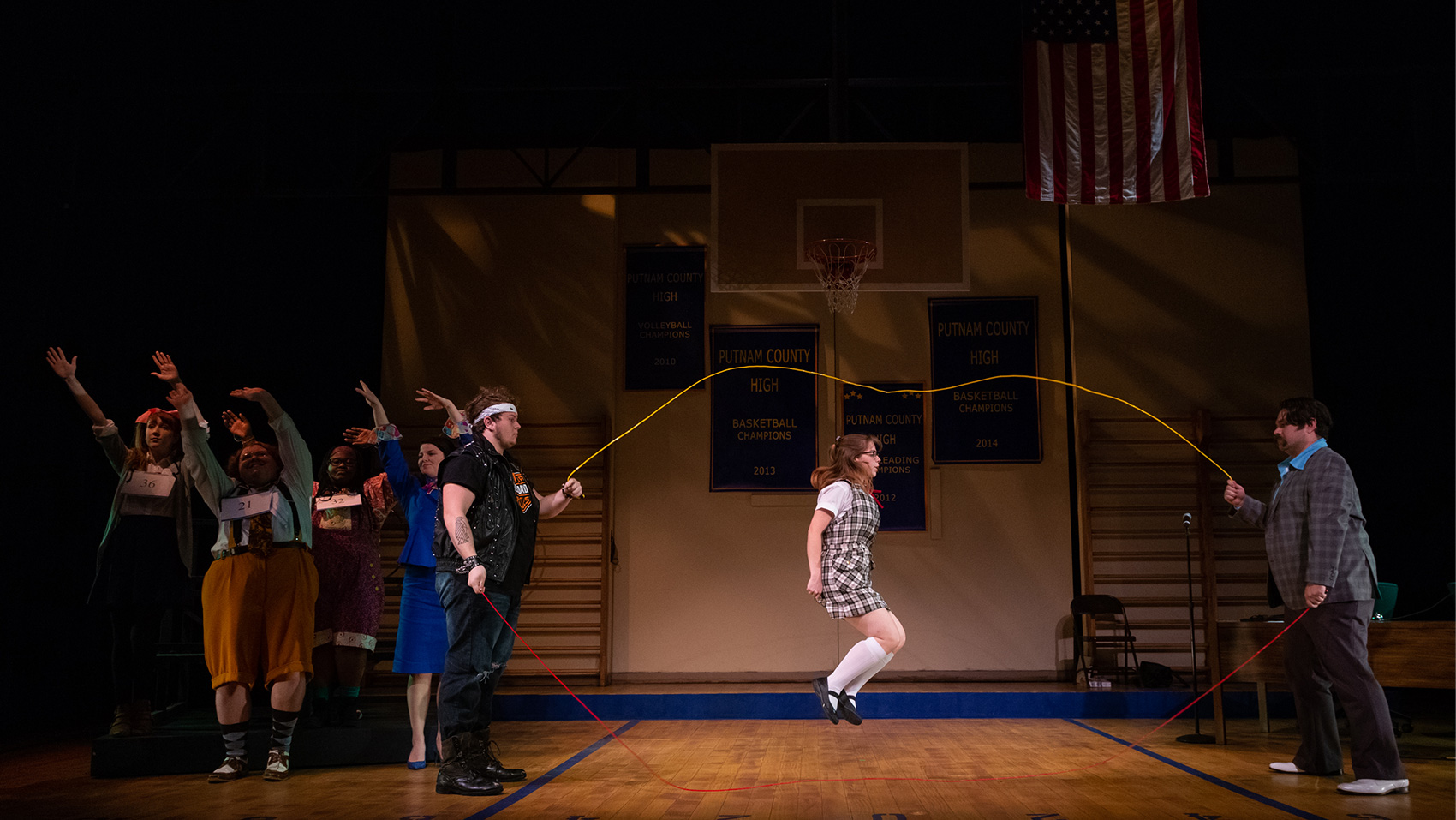 One of the contestants jumps ropes, as two men stand at either side holding the ropes. The other contestants sway along to the music in the background with their arms raised above their heads.