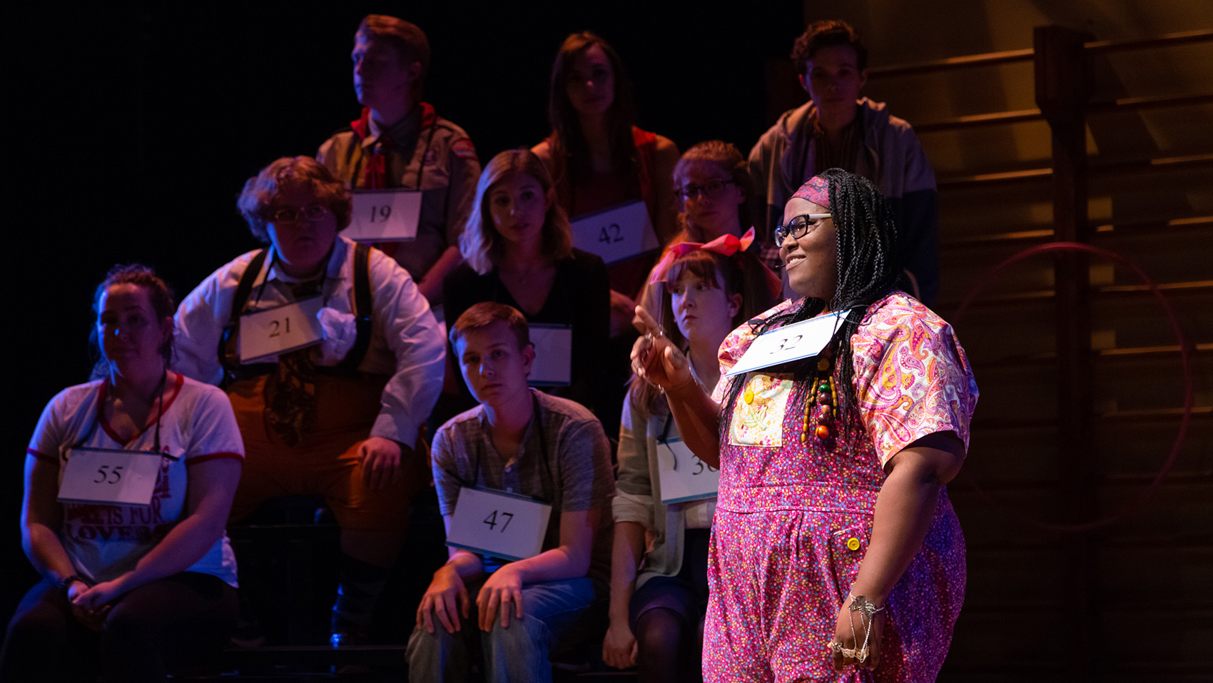 One of the spelling bee contestants sings under the spotlight as her fellow contestants watch from behind on the bleachers.