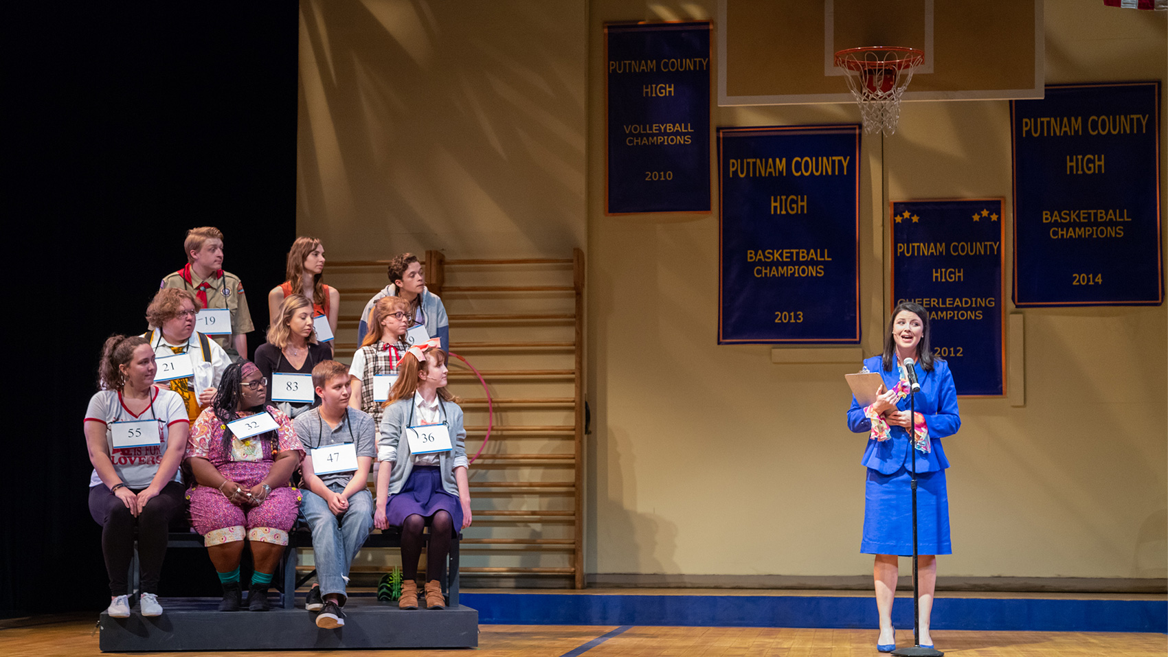 The host for the spelling bee contest stands in the middle of the stage (designed to look like a school gym with a basketball hoop mounted at the top and assorted wall hangings saying "Putnam County High") and speaks through a microphone. The contestants sit on bleachers to the side.