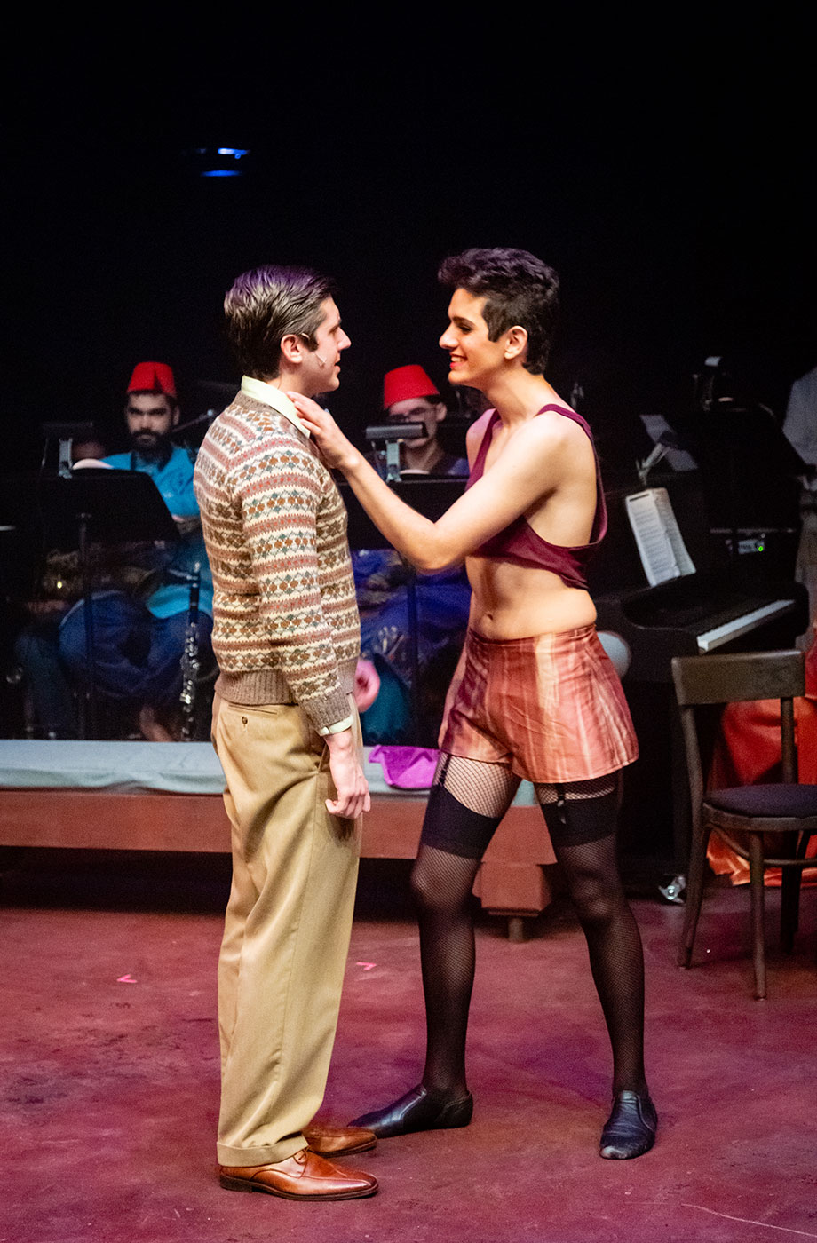 A male cabaret performer approaches another man, he puts his hand on the other man's shoulder. 