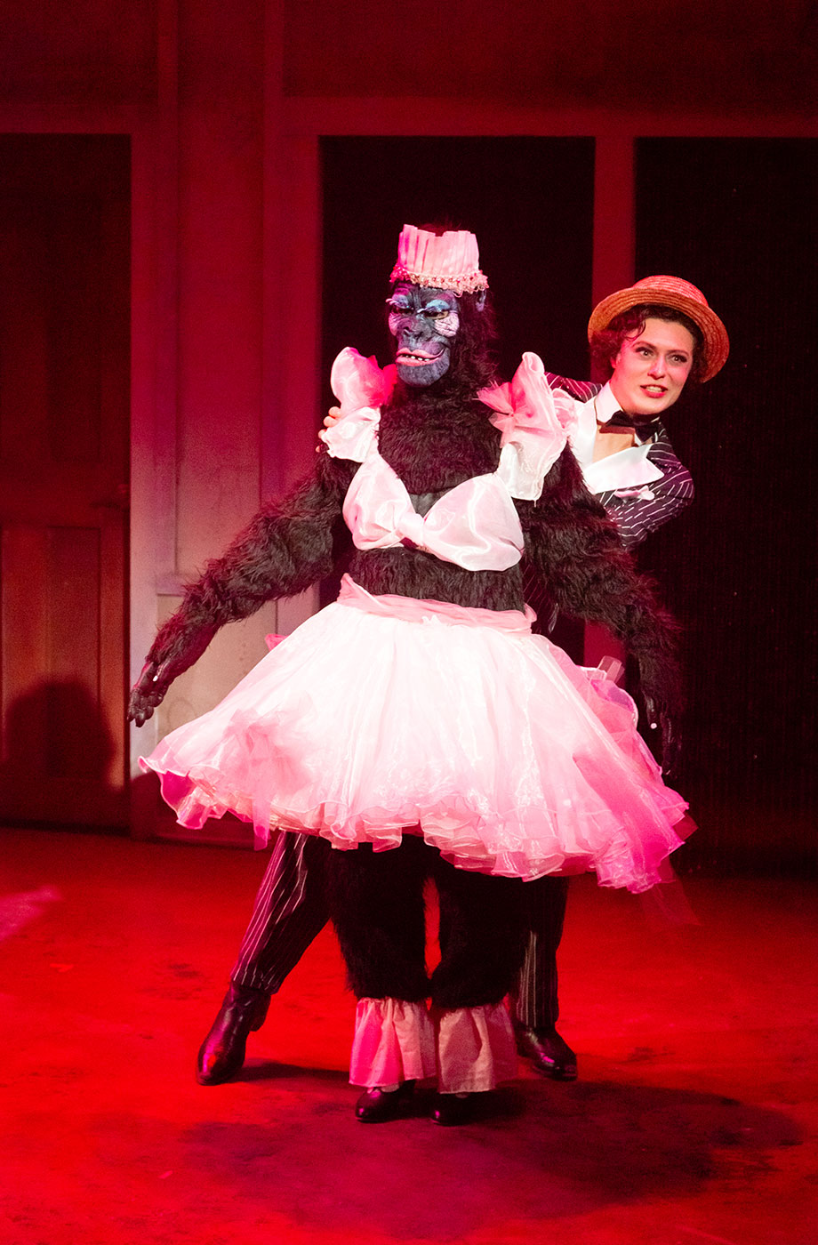 A male cabaret performer sticks his head out from behind a performer dressed in a gorilla suit wearing a tulle skirt and bra.