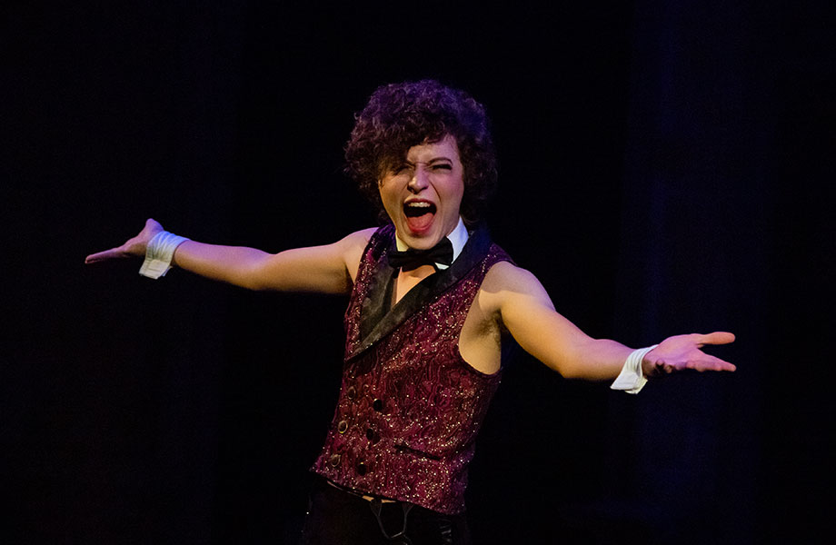 A male cabaret performer wearing a vest, bow tie, and cuffs sings,  sings enthusiastically. 