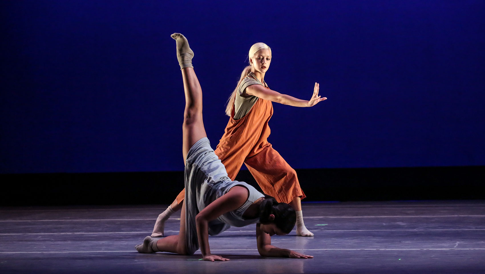 Two women dancers on stage, one supports herself on the ground with her arms and extends her leg straight upward.