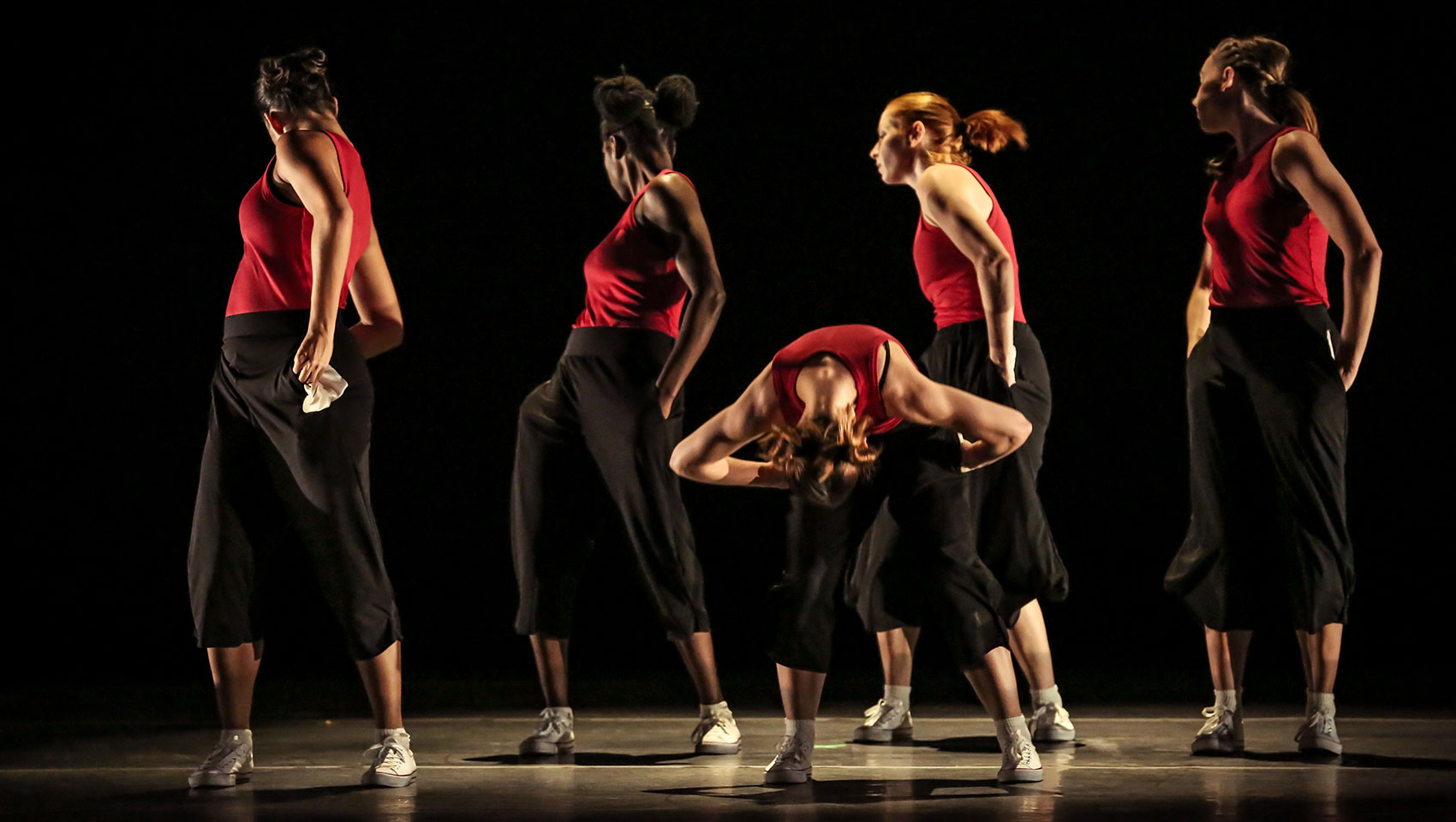 Five female dancers. Four in the back all face backwards, the one in the front is bent forward at her waist. 