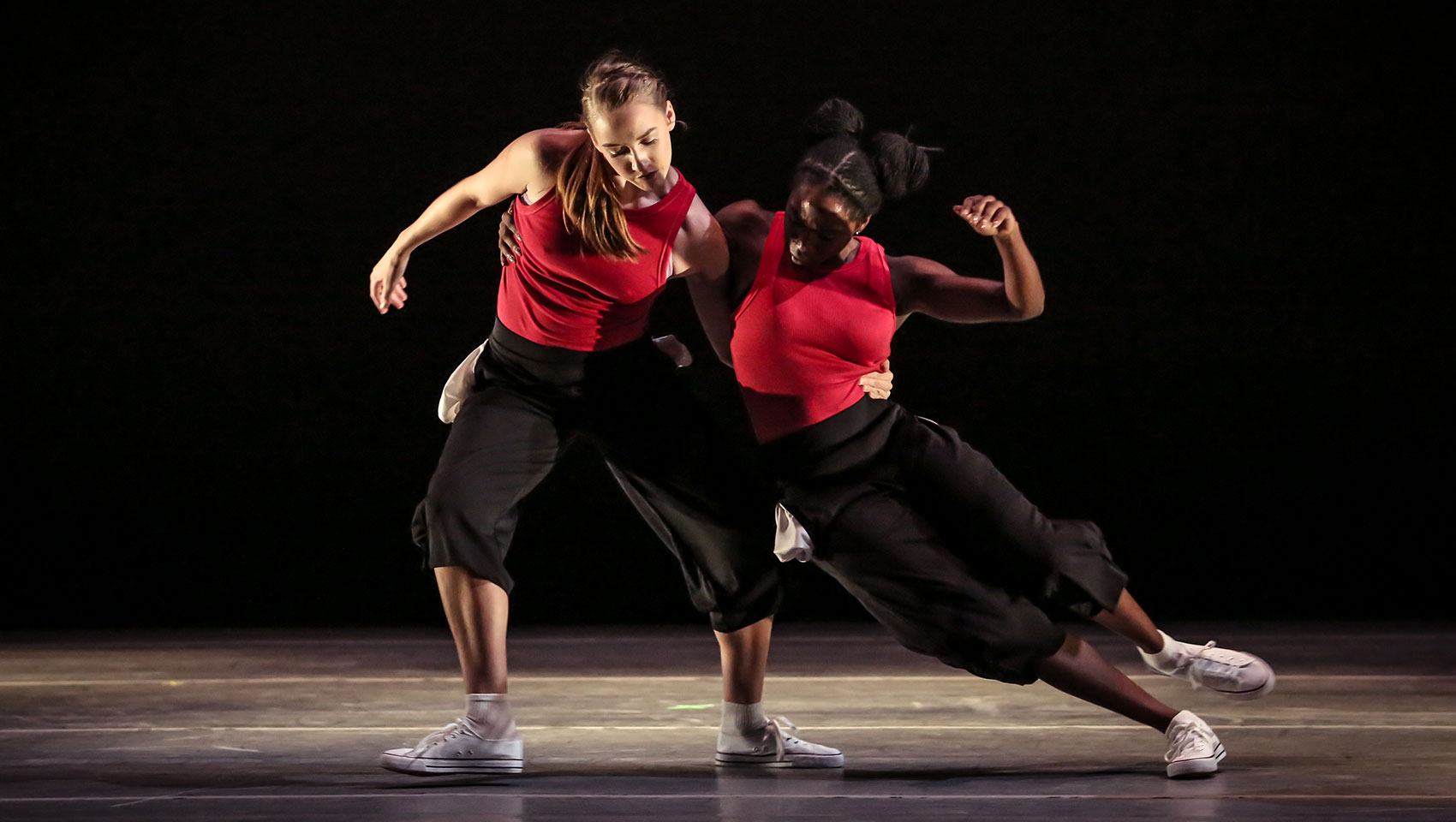 A female dancer supports another female dancer with her arm around her waist as she takes a turn around the floor.