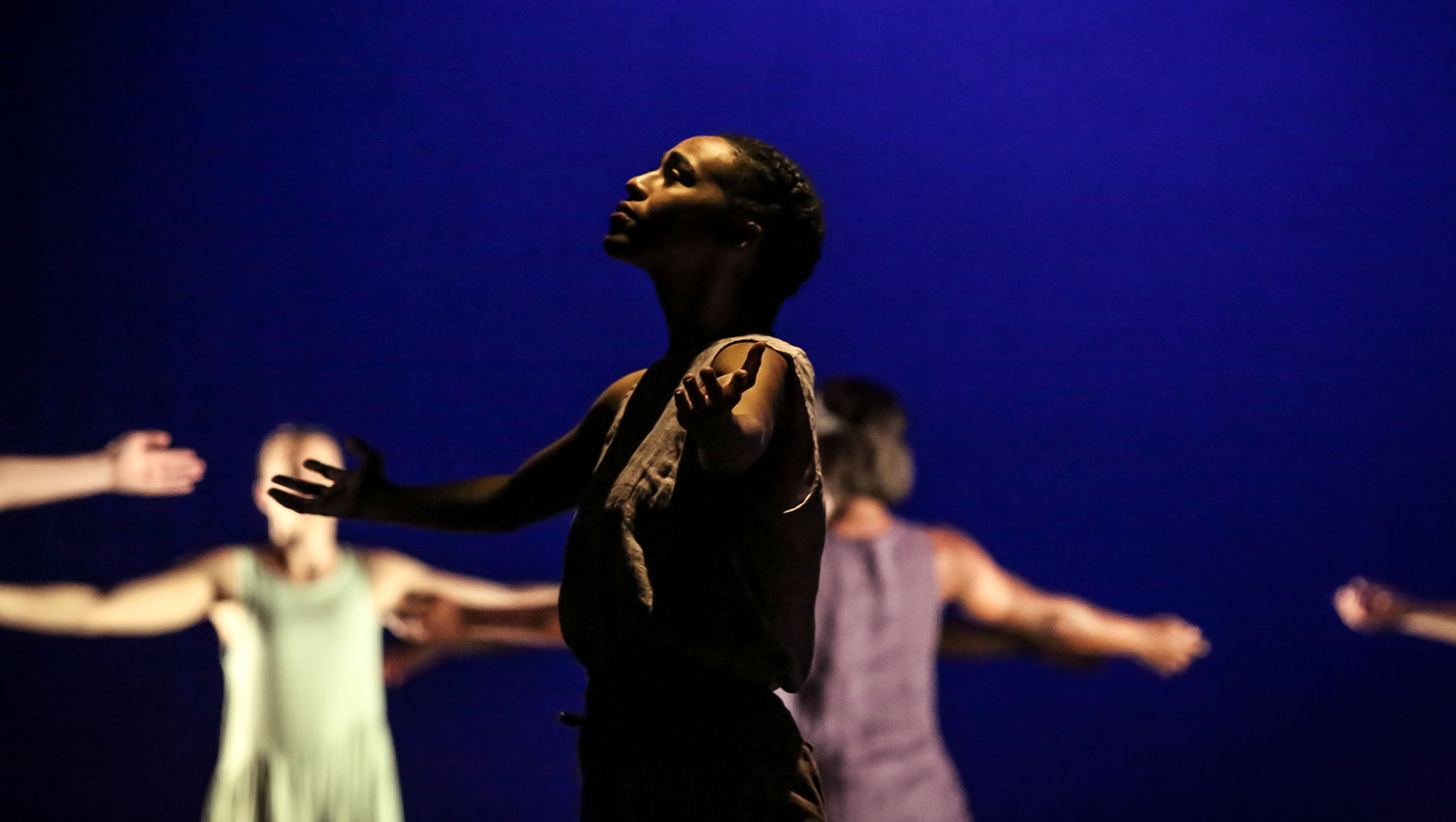 A female dancer, her face in shadow, holds out her arms and raises her face upwards.