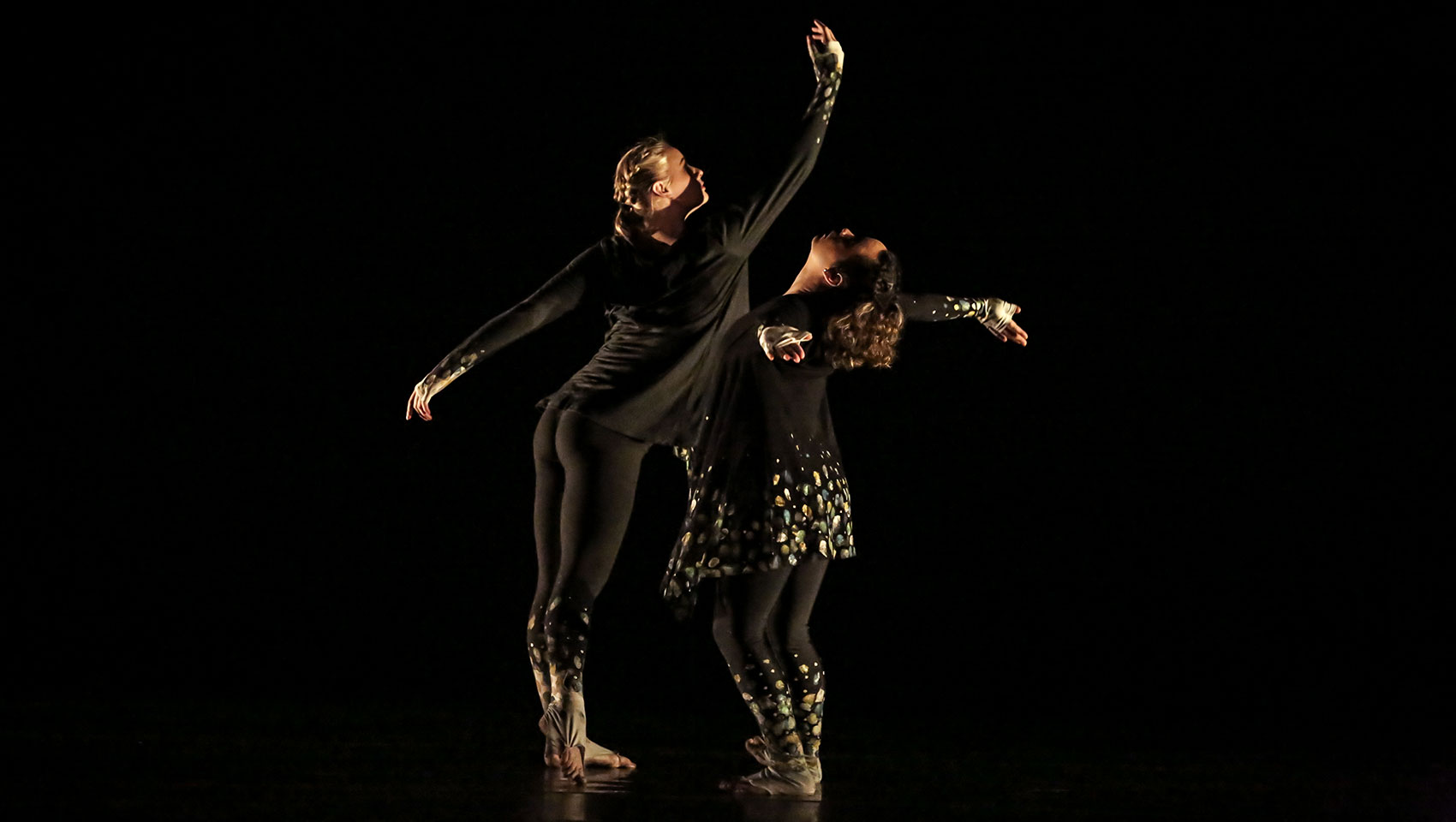 Two female dancers dressed all in black extend their arms outwards.