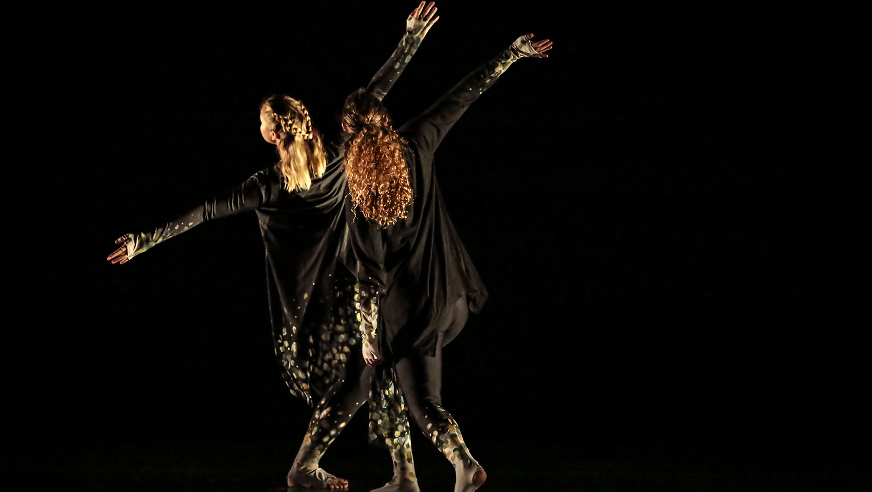 Two female dancers dressed in all black extend their arms outwards and bend their knees slightly, leaning to their sides. 