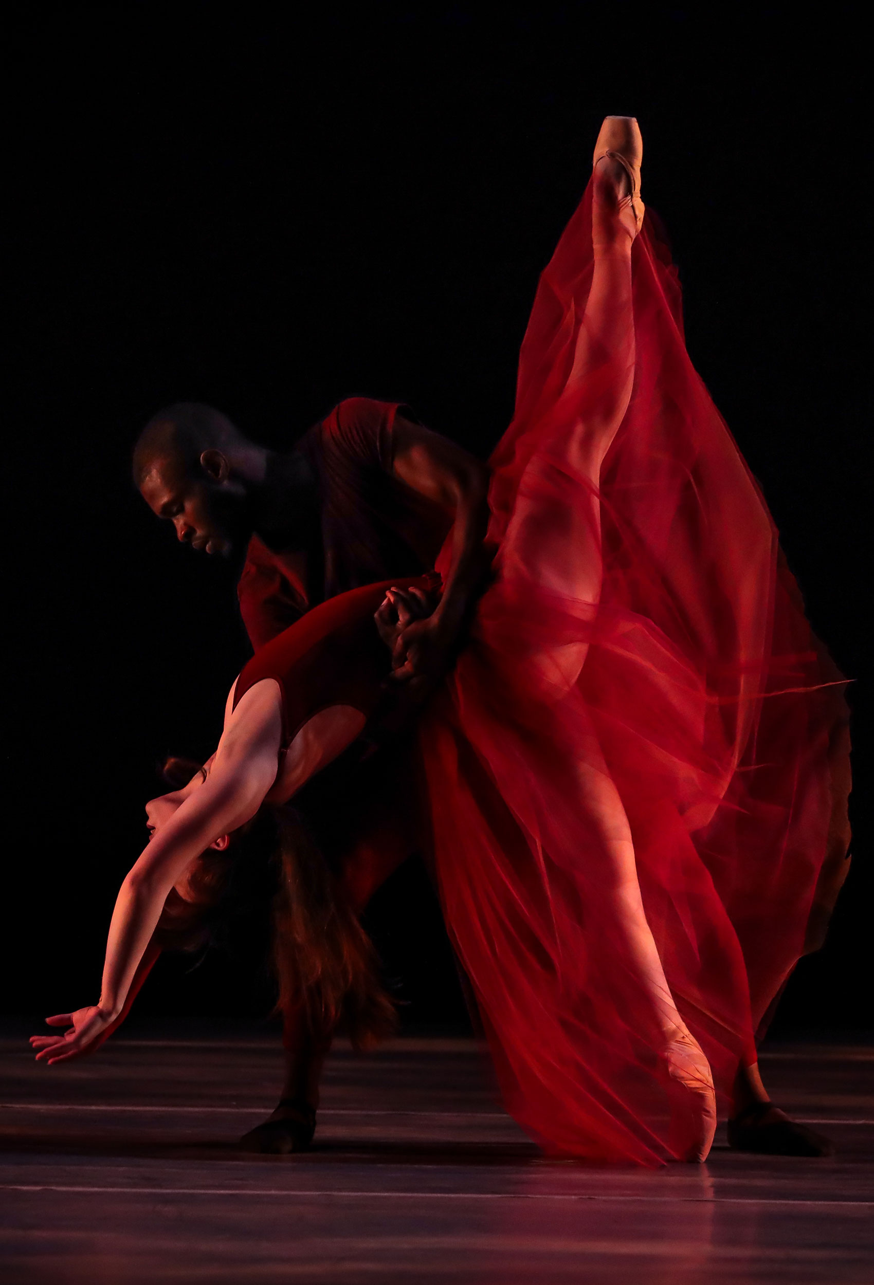 A Black male dancer holds a female dancer in his arms. He leans forward, holding her as she extends her arms gracefully backwards toward the floor and extends one leg straight upwards. 