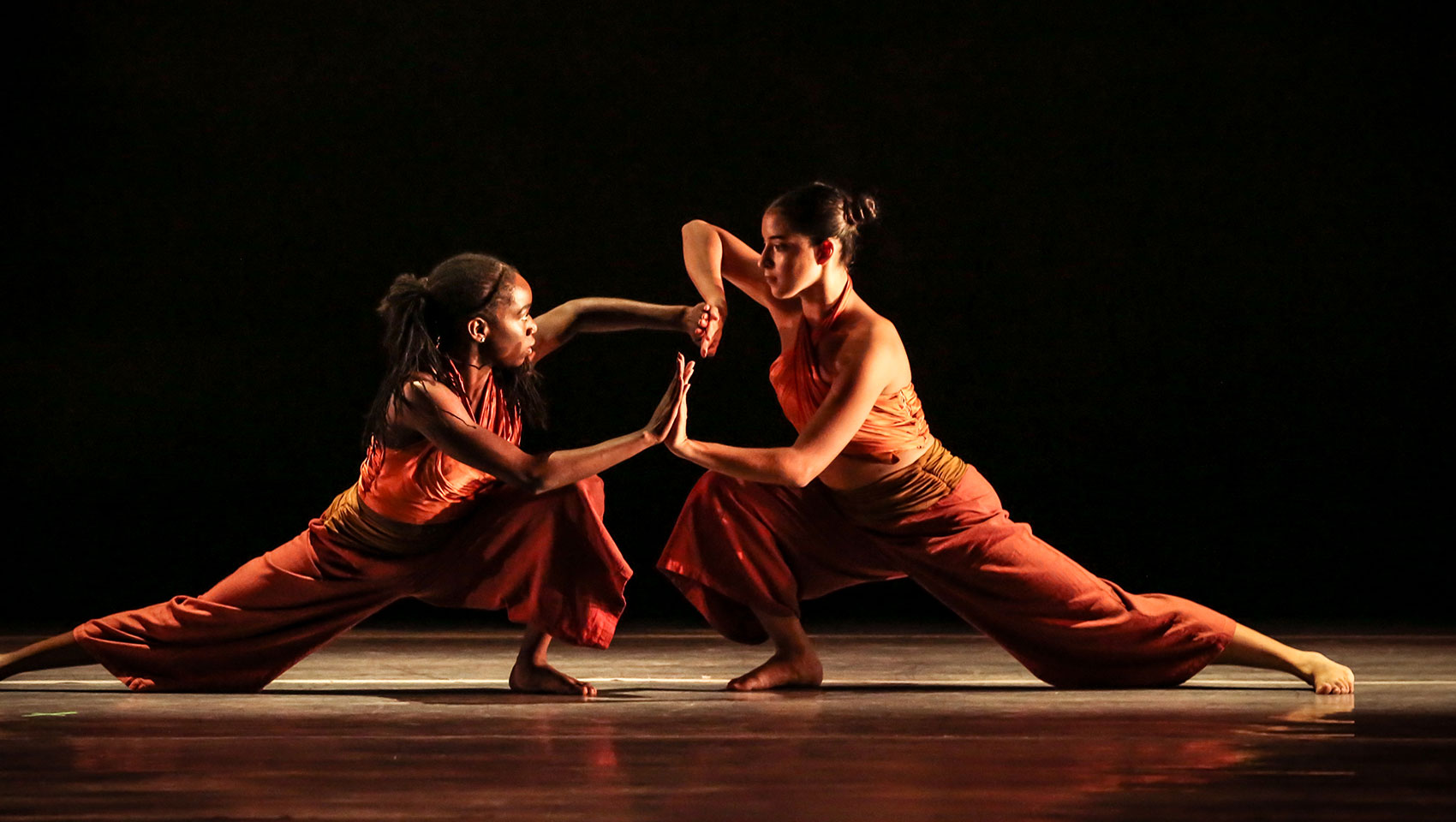 Two female dancers, one Black and one Latina, lunge forward on the ground facing each other. They meet their palms and hold each other's gaze.