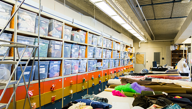 Storage bins containing multiple fabrics and assorted costume-making materials are lined in shelves along one wall of the costume shop. A dress form stands among the tables covered with fabrics and tools. 