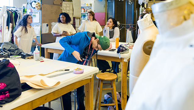Students working in the costume shop. One student closely examines the costume laid out on the table, he's holding a pair of scissors in one hand.