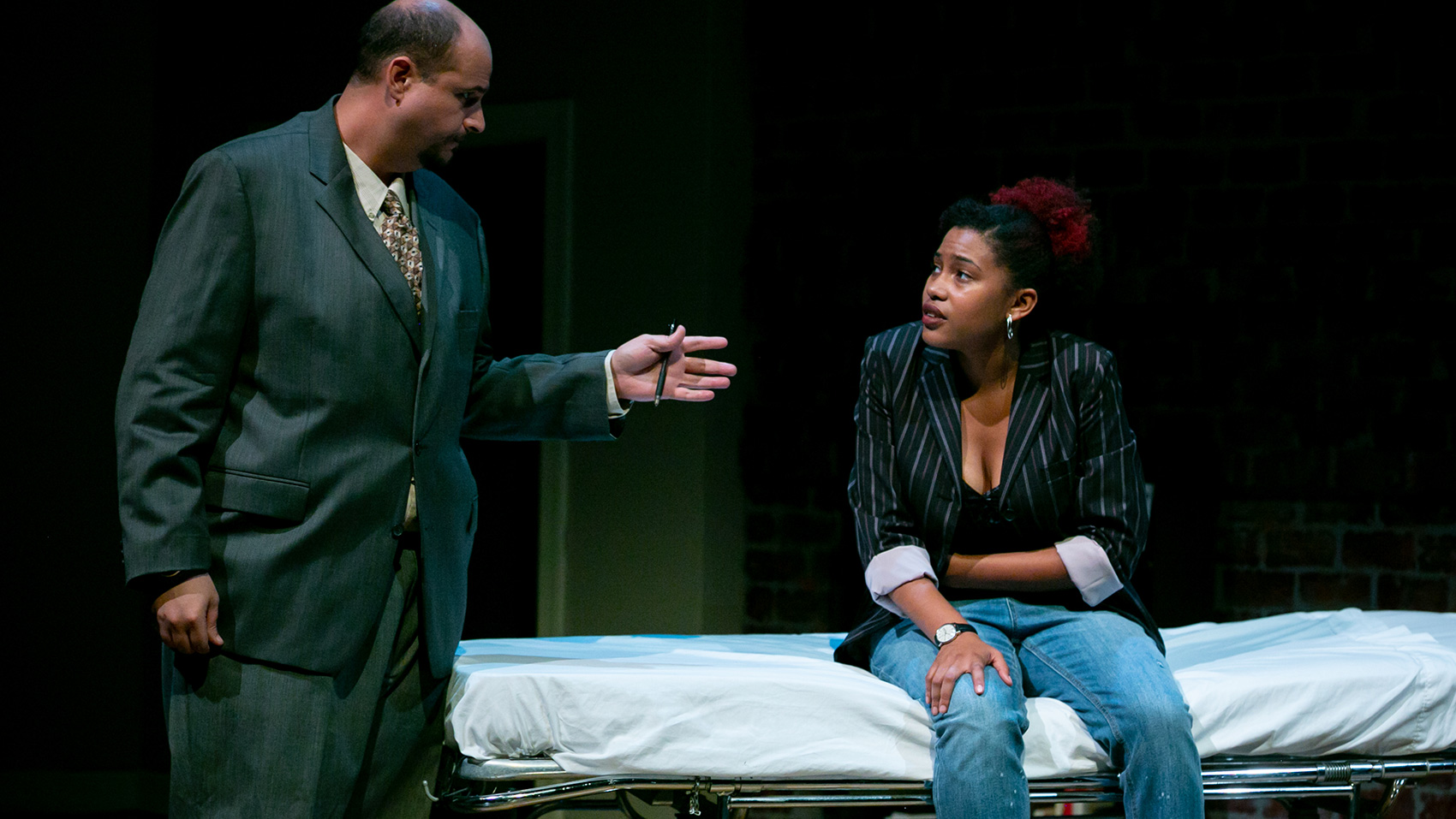 A man wearing a suit talks to one of the main women characters, who is sitting on a gurney.