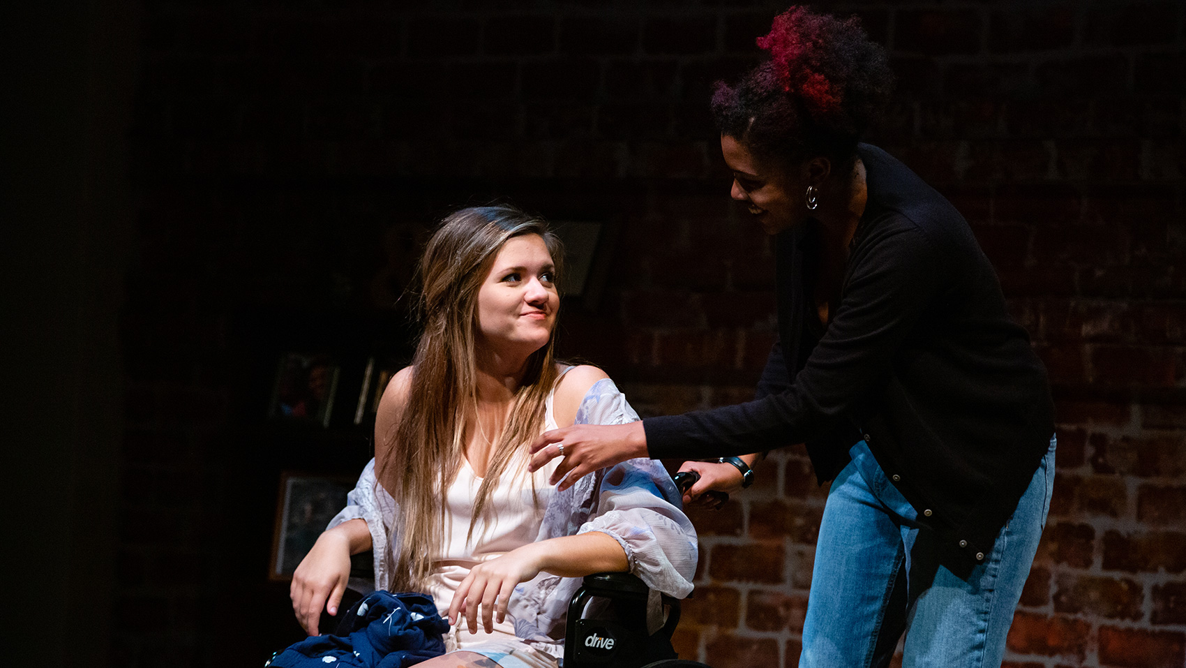 A woman slightly bending forward to speak with another woman who is using a wheelchair.