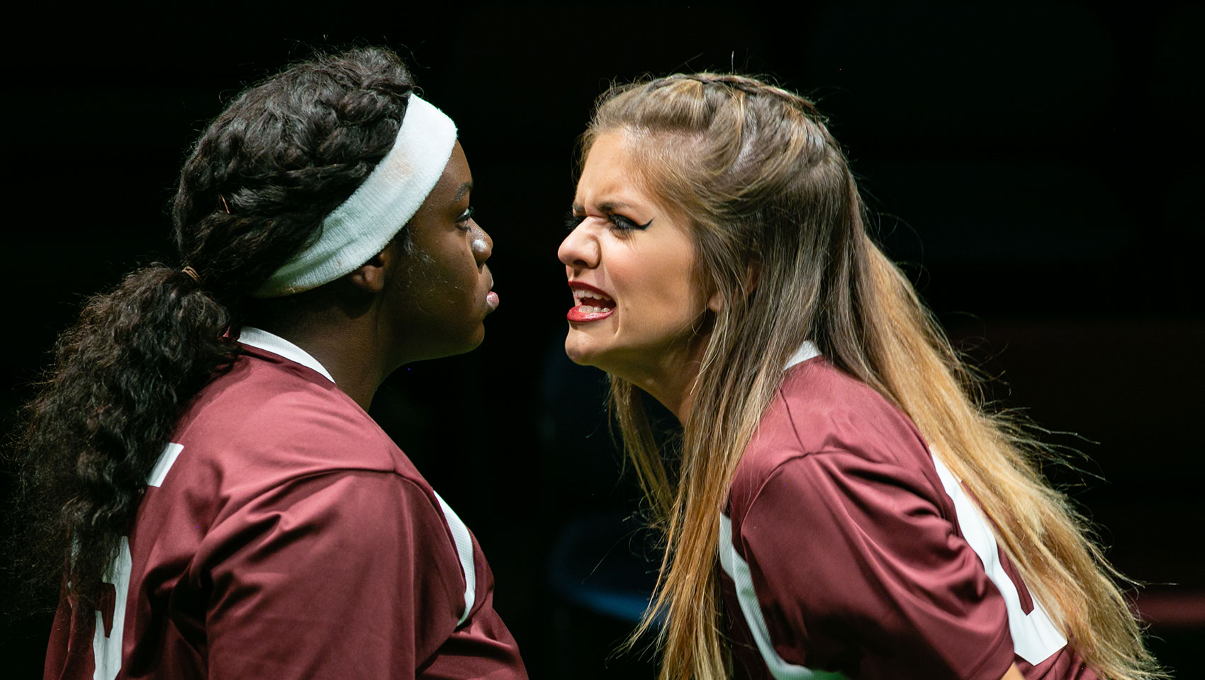 A tense scene between two members of the same girls' scocer team. One of them, a Black girl, stares ahead stone-faced, while the other, a white girl, gets in her  face with a grimacing expression. 