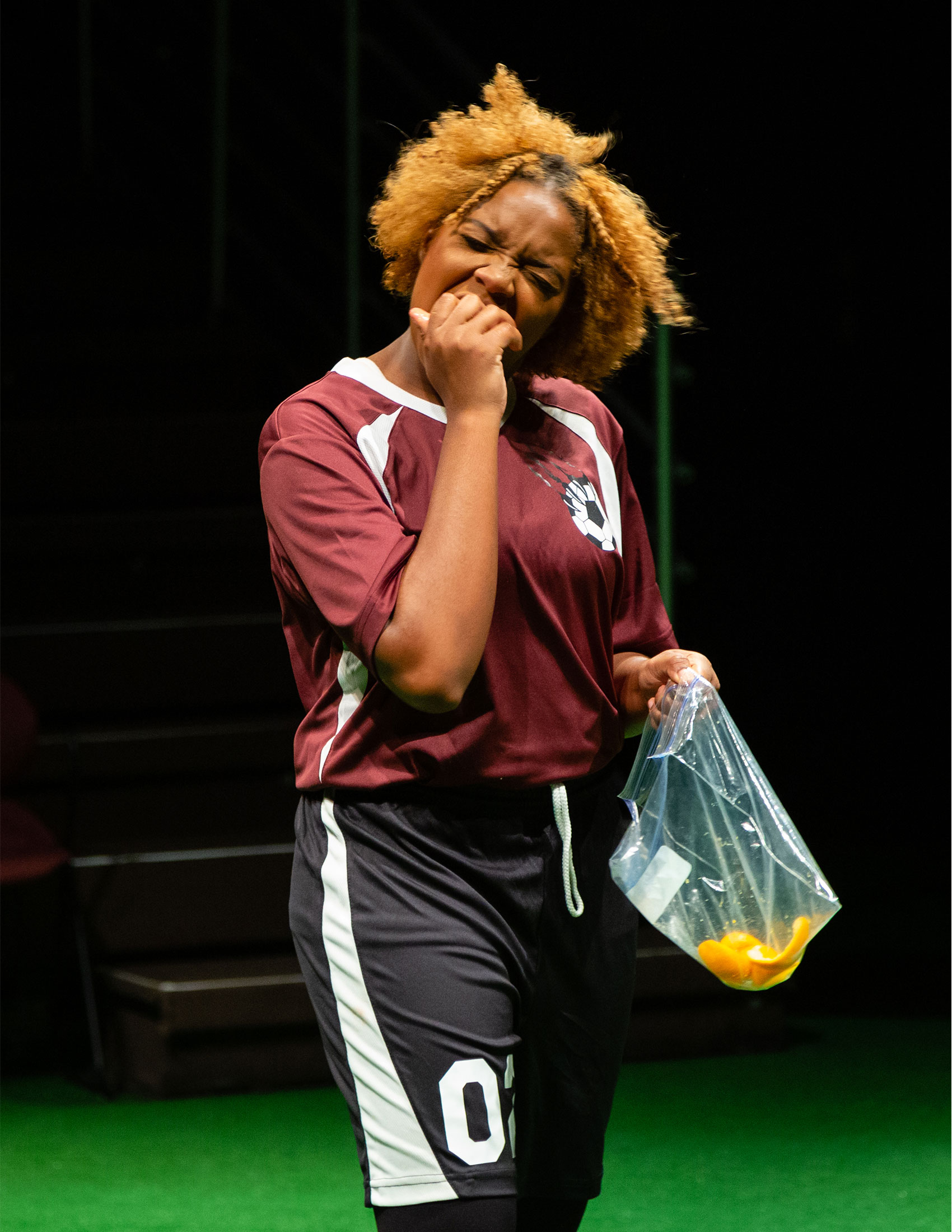 A Black girl wearing a soccer uniform eats an orange with one hand, in the other she holds a ziplock bag containing orange peels.