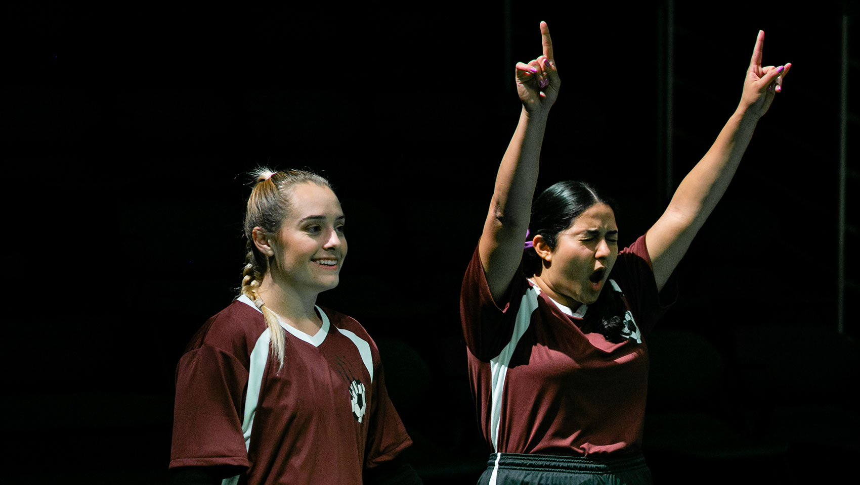 Two women's soccer teammates stand near each other, one cheering as she raises both arms upwards in victory signs, the other smiling as she looks on whatever is happening before them. 